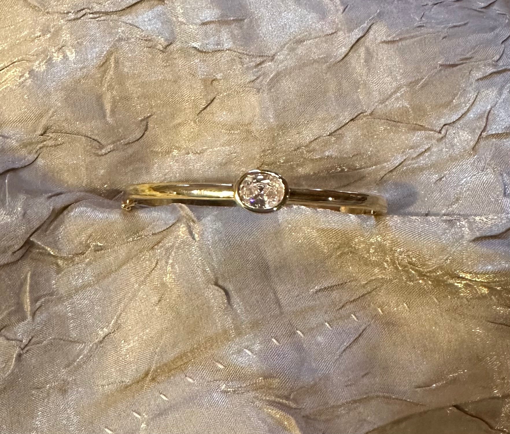 A gold-toned bangle bracelet with a single round clear gemstone, displayed on a textured, shimmering champagne-colored fabric background.
