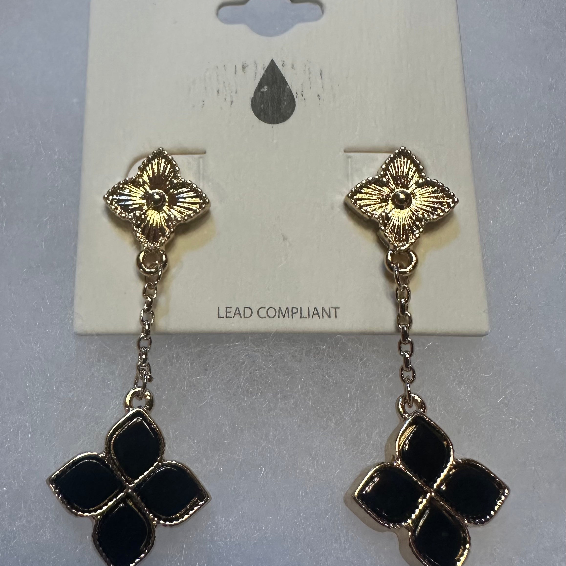 A pair of gold-tone dangle earrings with black enamel clover motifs, displayed on a white card backing labeled 'LEAD COMPLIANT'.