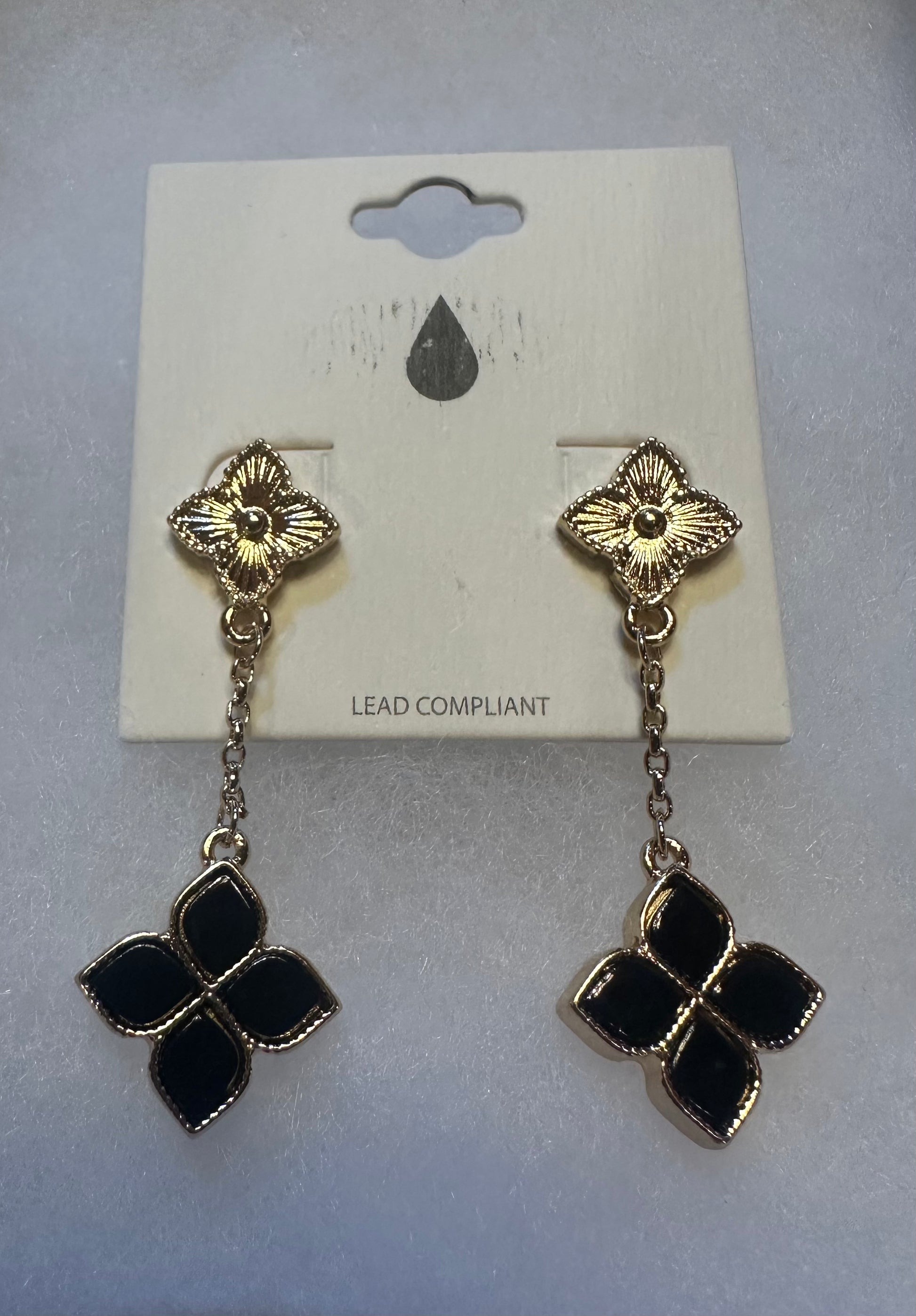 A pair of gold-tone dangle earrings with black enamel clover motifs, displayed on a white card backing labeled 'LEAD COMPLIANT'.