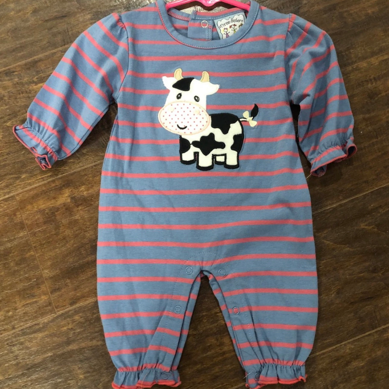 A baby's long-sleeve footless onesie with blue and pink horizontal stripes, featuring a black and white cow applique on the chest, laid flat on a dark wood floor.
