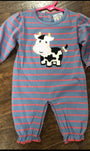 A baby's long-sleeve footless onesie with blue and pink horizontal stripes, featuring a black and white cow applique on the chest, laid flat on a dark wood floor.