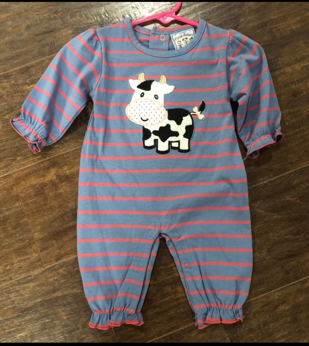 A baby's long-sleeve footless onesie with blue and pink horizontal stripes, featuring a black and white cow applique on the chest, laid flat on a dark wood floor.