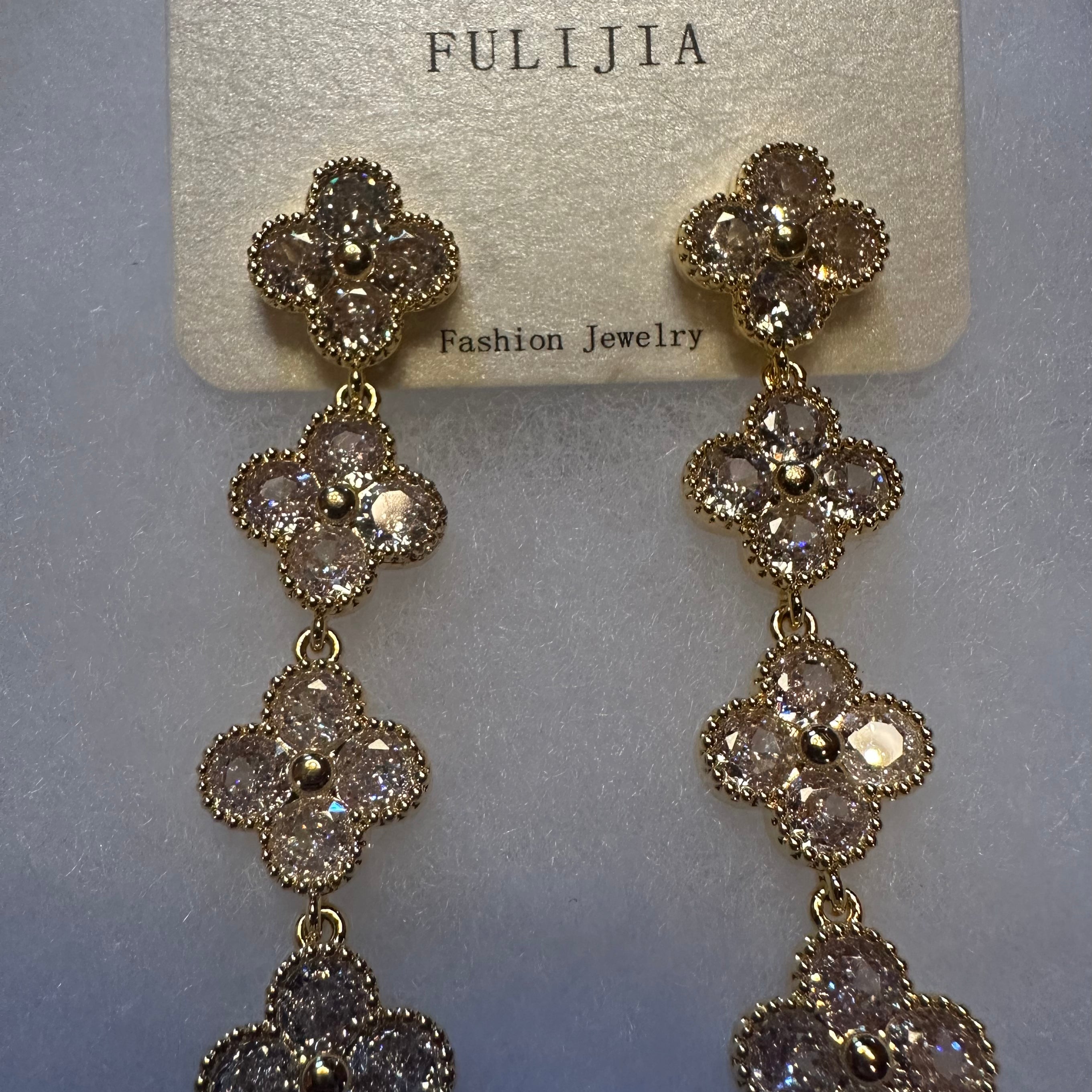 A pair of gold-tone dangle earrings with three floral clover motifs, each adorned with clear rhinestones, hanging from a textured beige card labeled 'FULIJIA'.