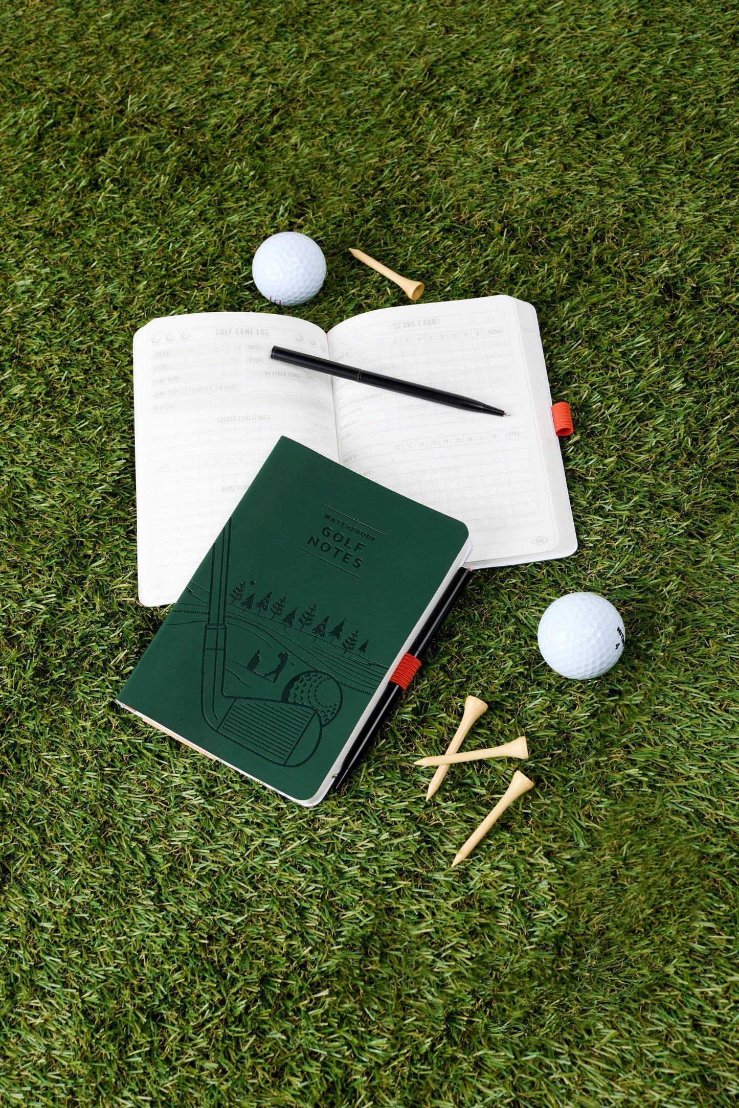 Open notebook with pen, green notebook, golf balls, and tees on grass