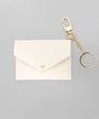 A small off-white envelope-style card holder or wallet with a gold snap button, attached to a gold keychain clip and ring, shown on a plain light gray background.