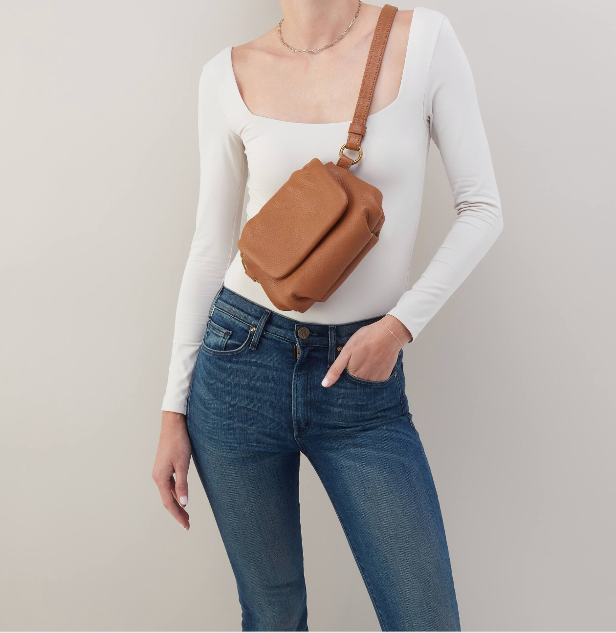Person wearing a white long-sleeve top, blue jeans, and carrying a brown crossbody bag on a plain background