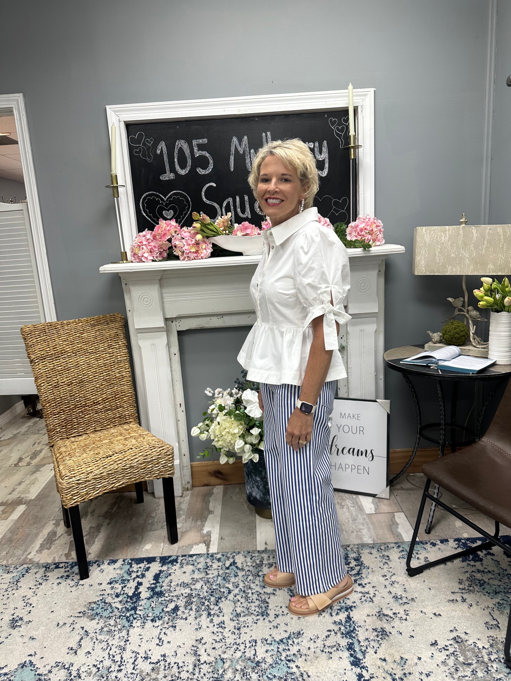 Woman standing in a room with decor elements like a fireplace, chair, and flowers.