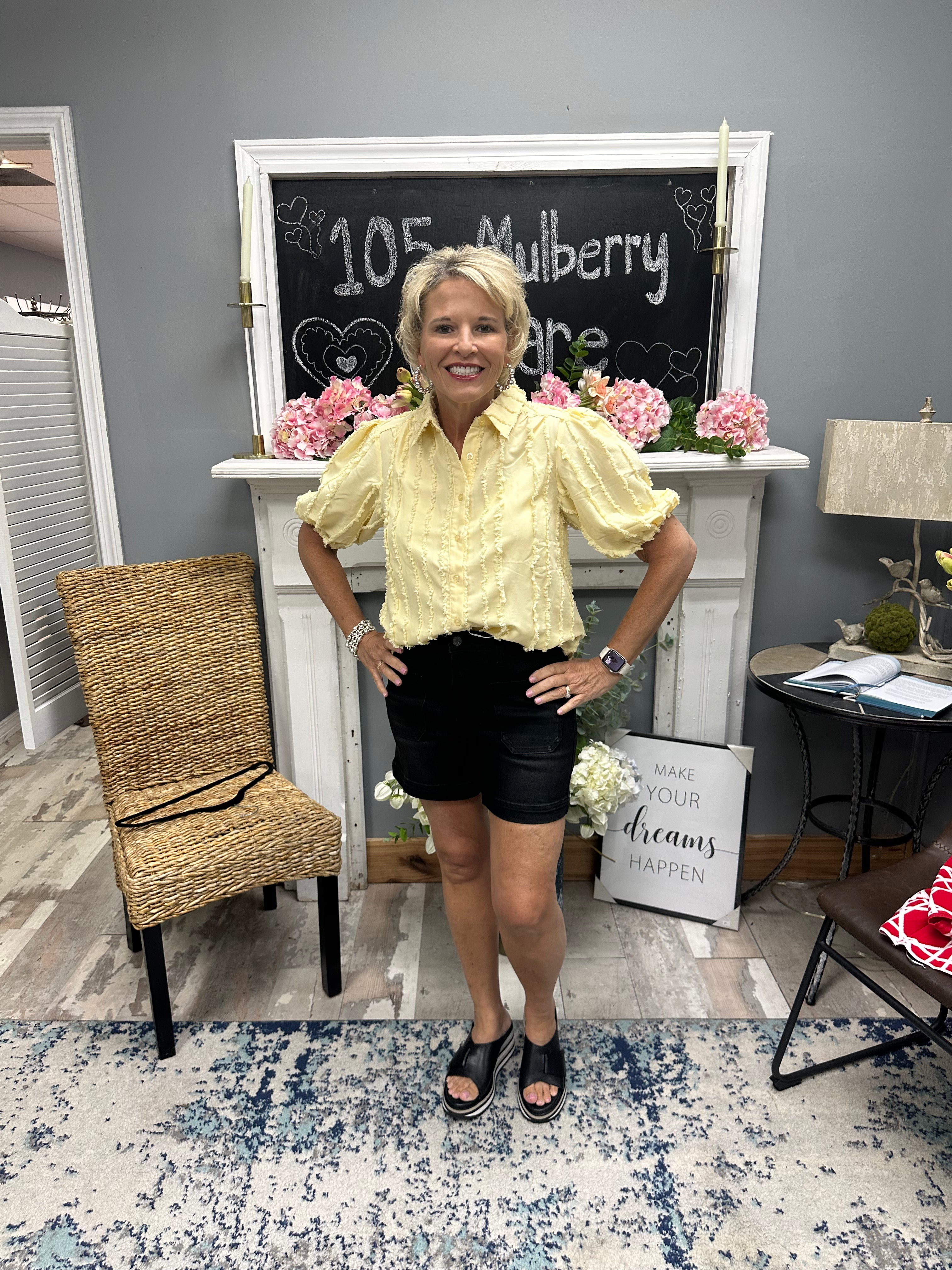 Woman in a yellow blouse and black shorts standing in a room with decor elements.