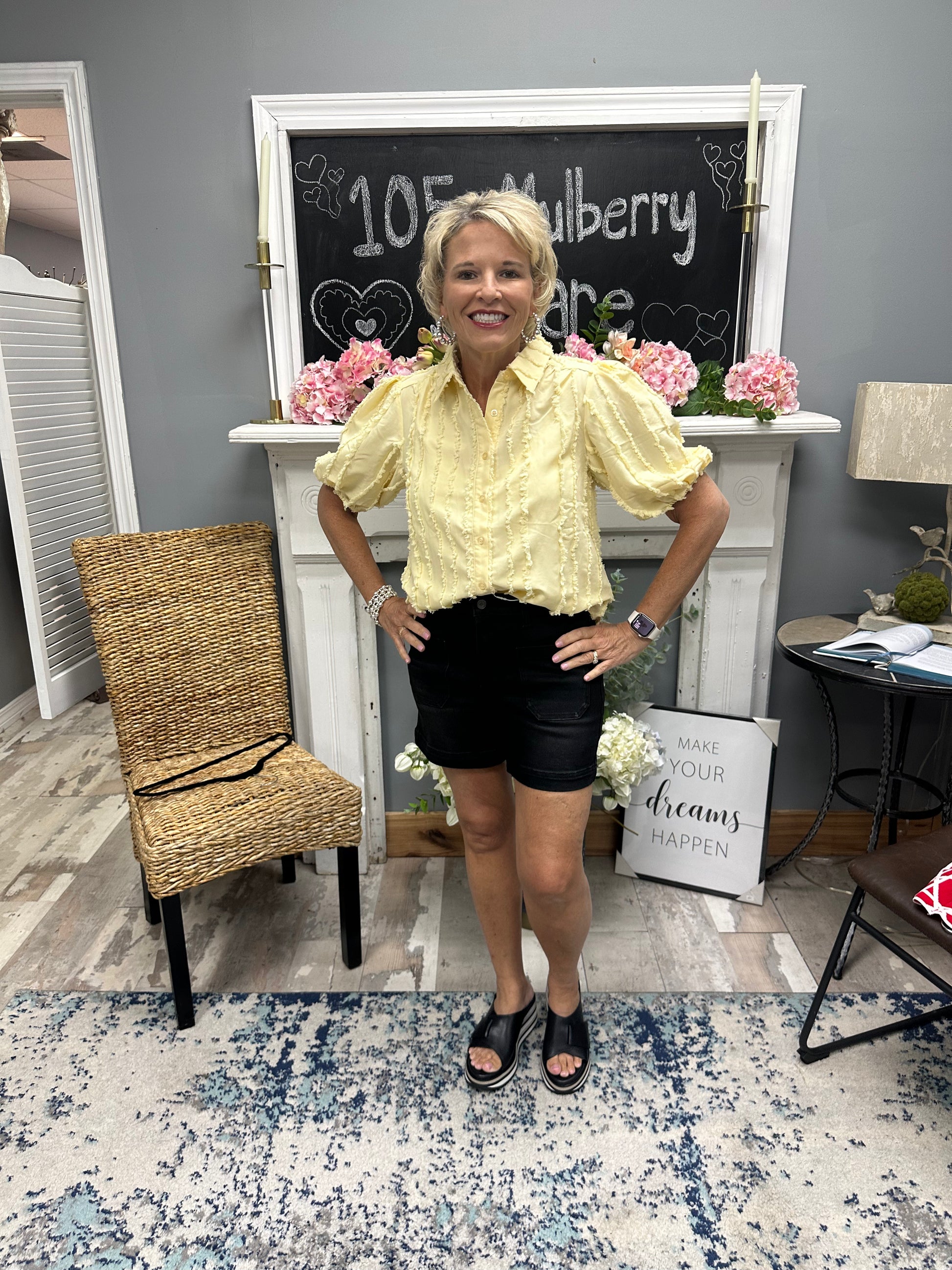 Woman in a yellow blouse and black shorts standing in a room with a fireplace and decor.