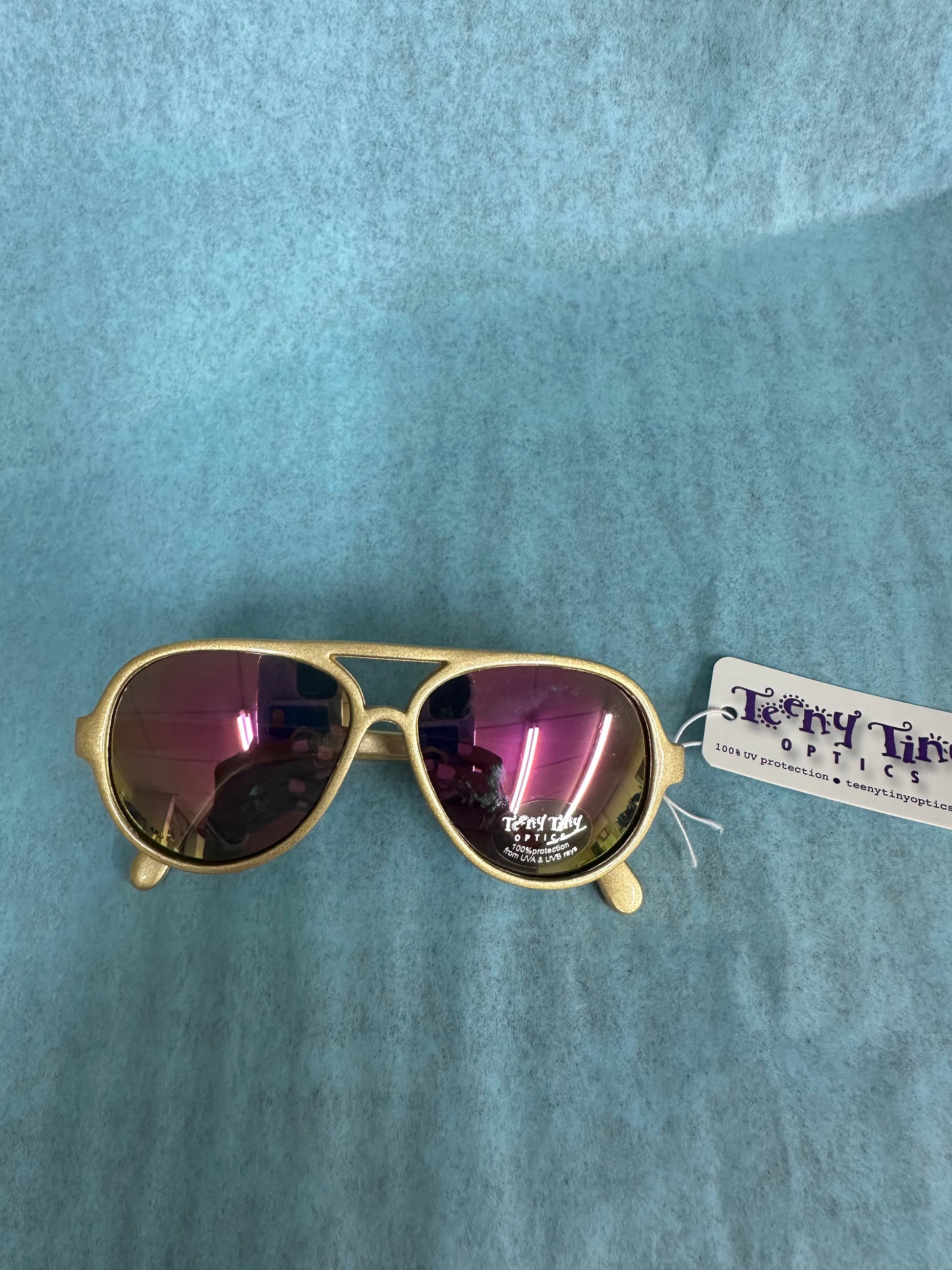 Gold aviator sunglasses with a purple lens on a blue textured background, featuring a tag from Tony Tun Optics.