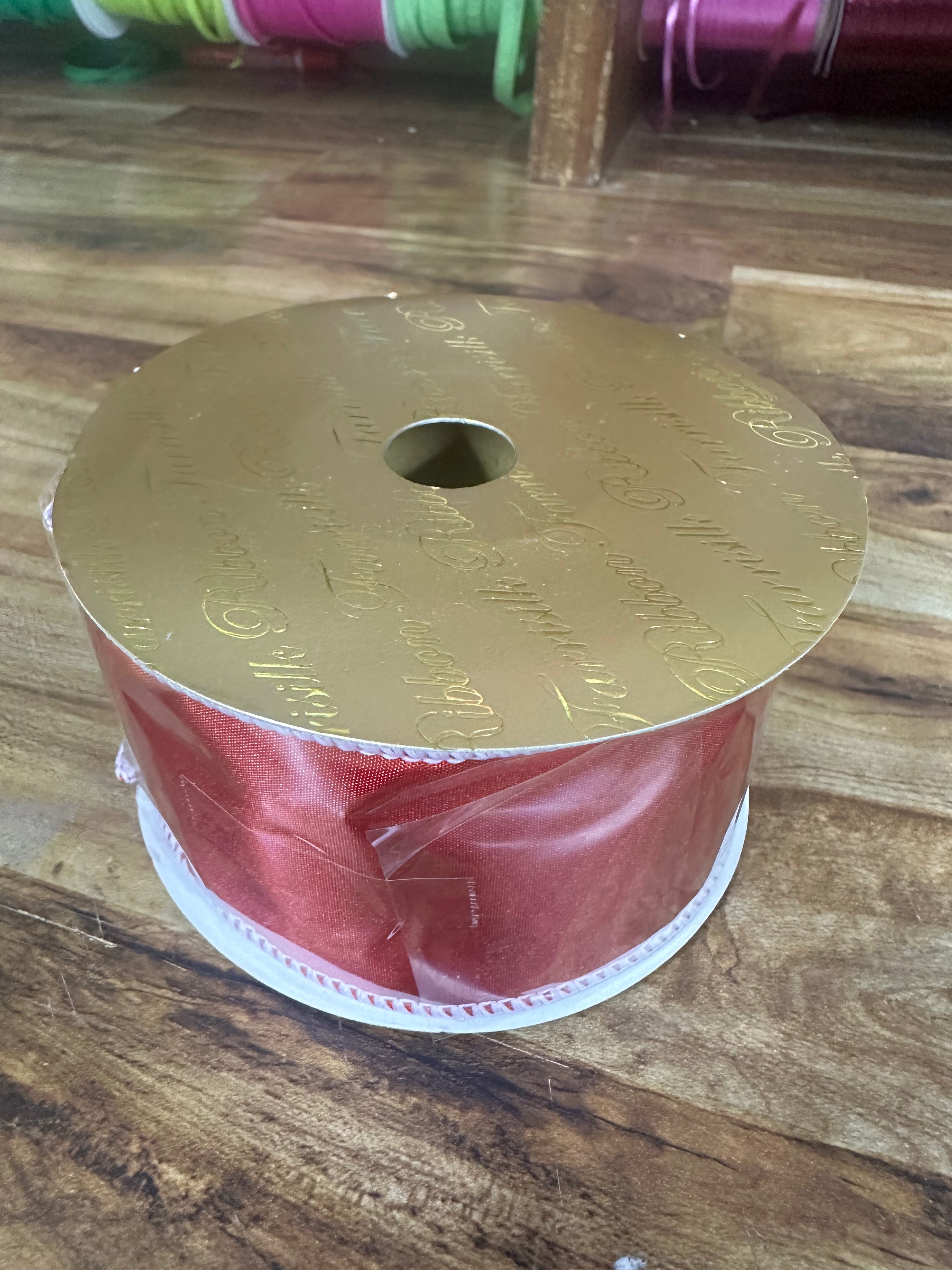 A spool of shiny red ribbon with a gold foil top and a white plastic base, placed on a wooden tabletop.