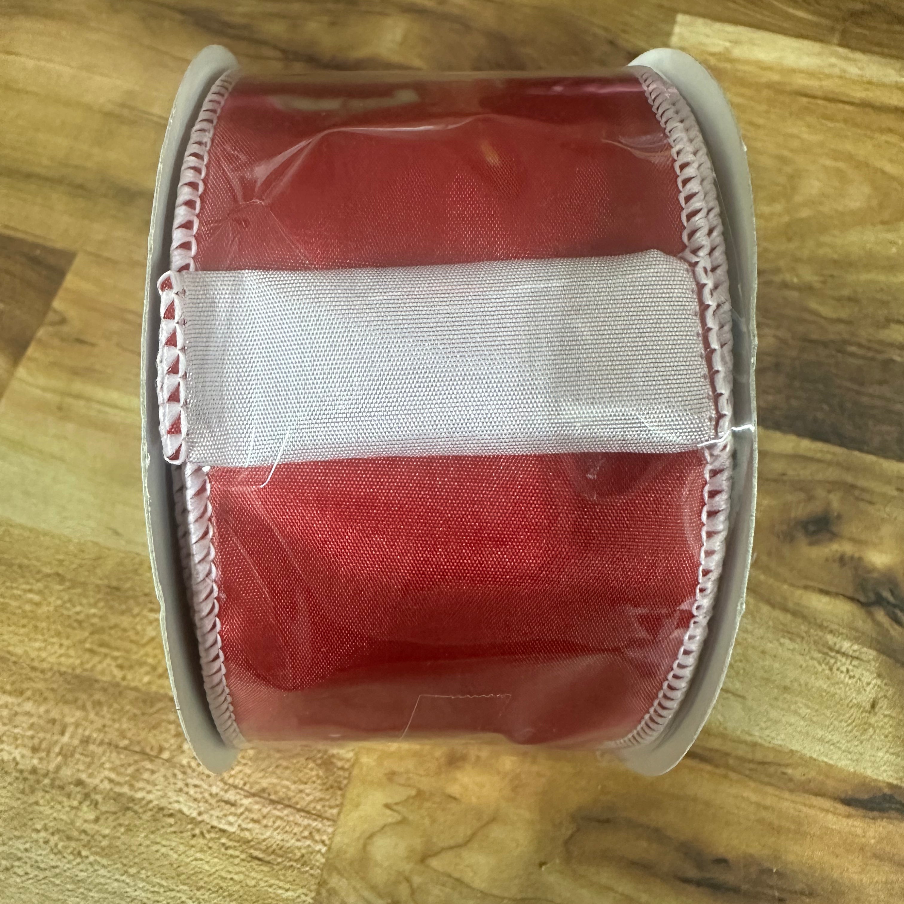 A roll of red grosgrain ribbon with a white center stripe and white stitched edges, wrapped in clear protective plastic, resting on a wooden surface.