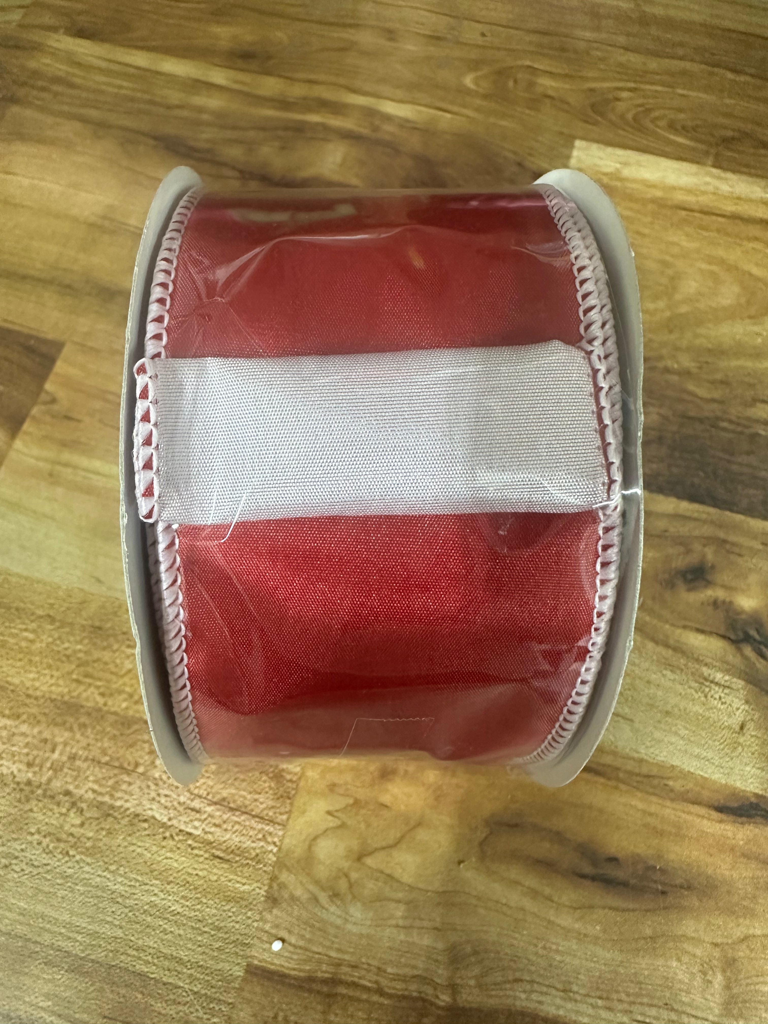 A roll of red grosgrain ribbon with a white center stripe and white stitched edges, wrapped in clear protective plastic, resting on a wooden surface.
