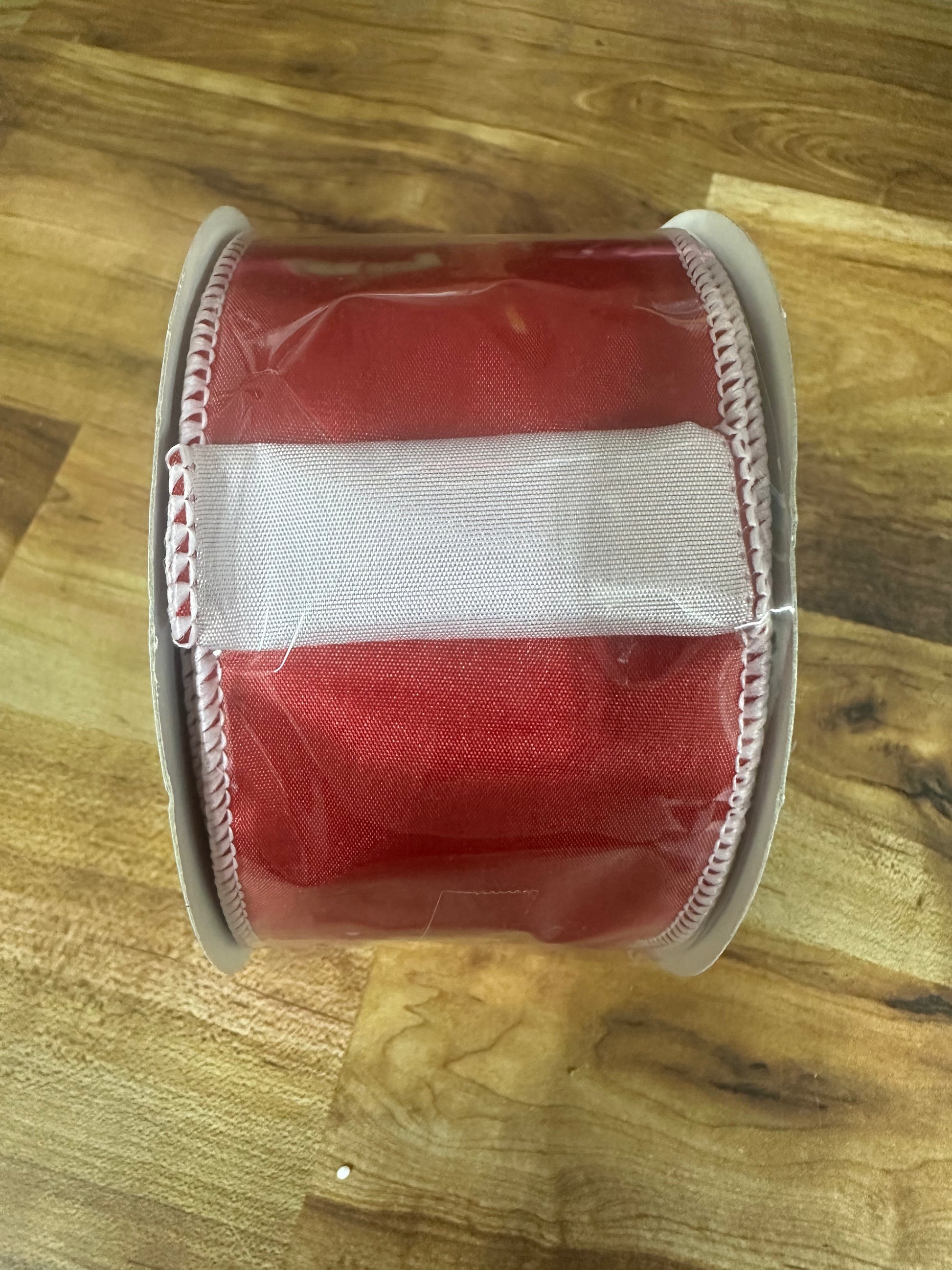 A roll of red grosgrain ribbon with a white center stripe and white stitched edges, wrapped in clear protective plastic, resting on a wooden surface.