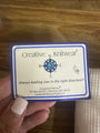 A hand holding a square white card with a blue border that reads 'Creative Knitwear' and a small compass logo, photographed over a wooden surface.