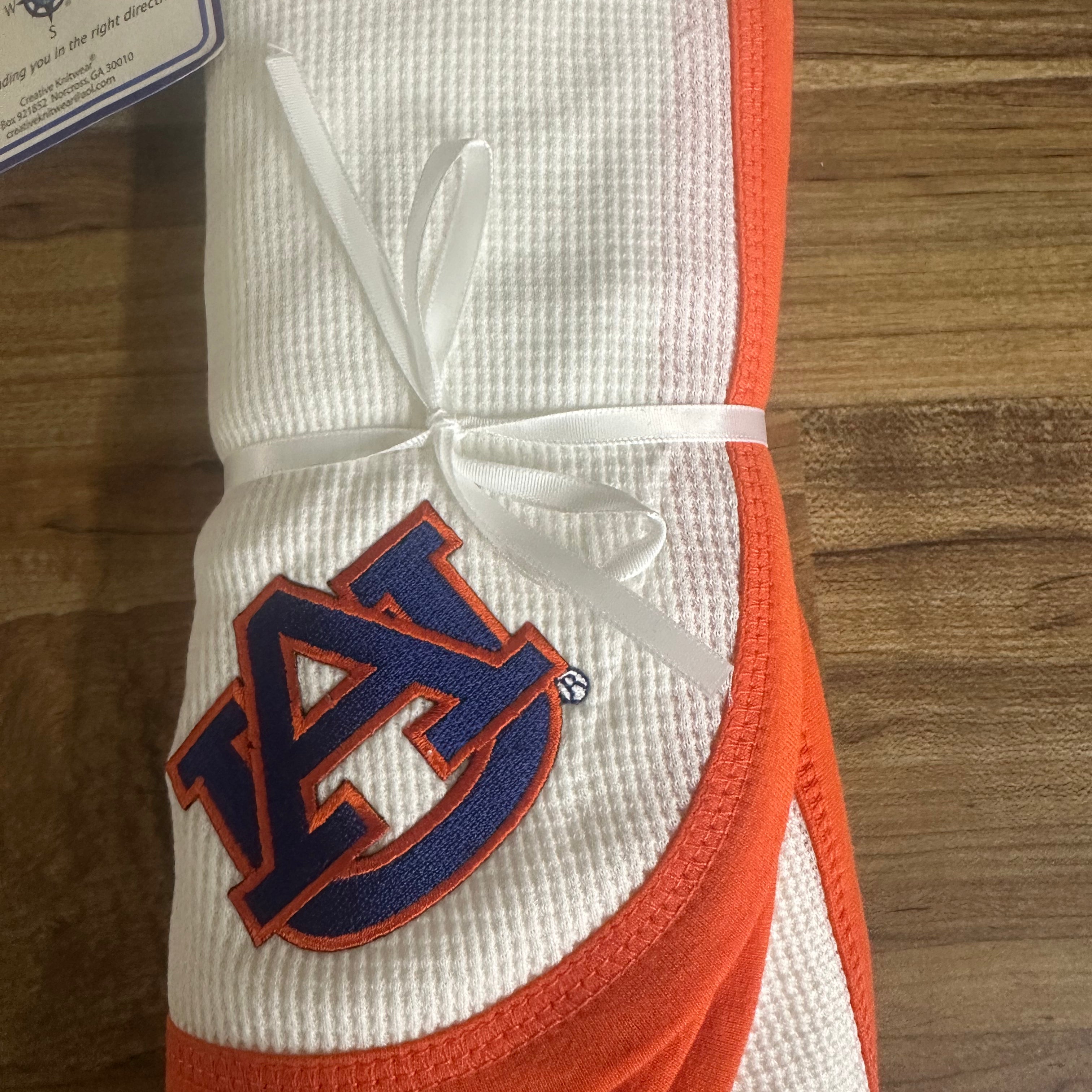 A folded white waffle-knit baby blanket with orange trim and a navy blue Auburn Tigers logo, tied with a white ribbon on a wooden surface.