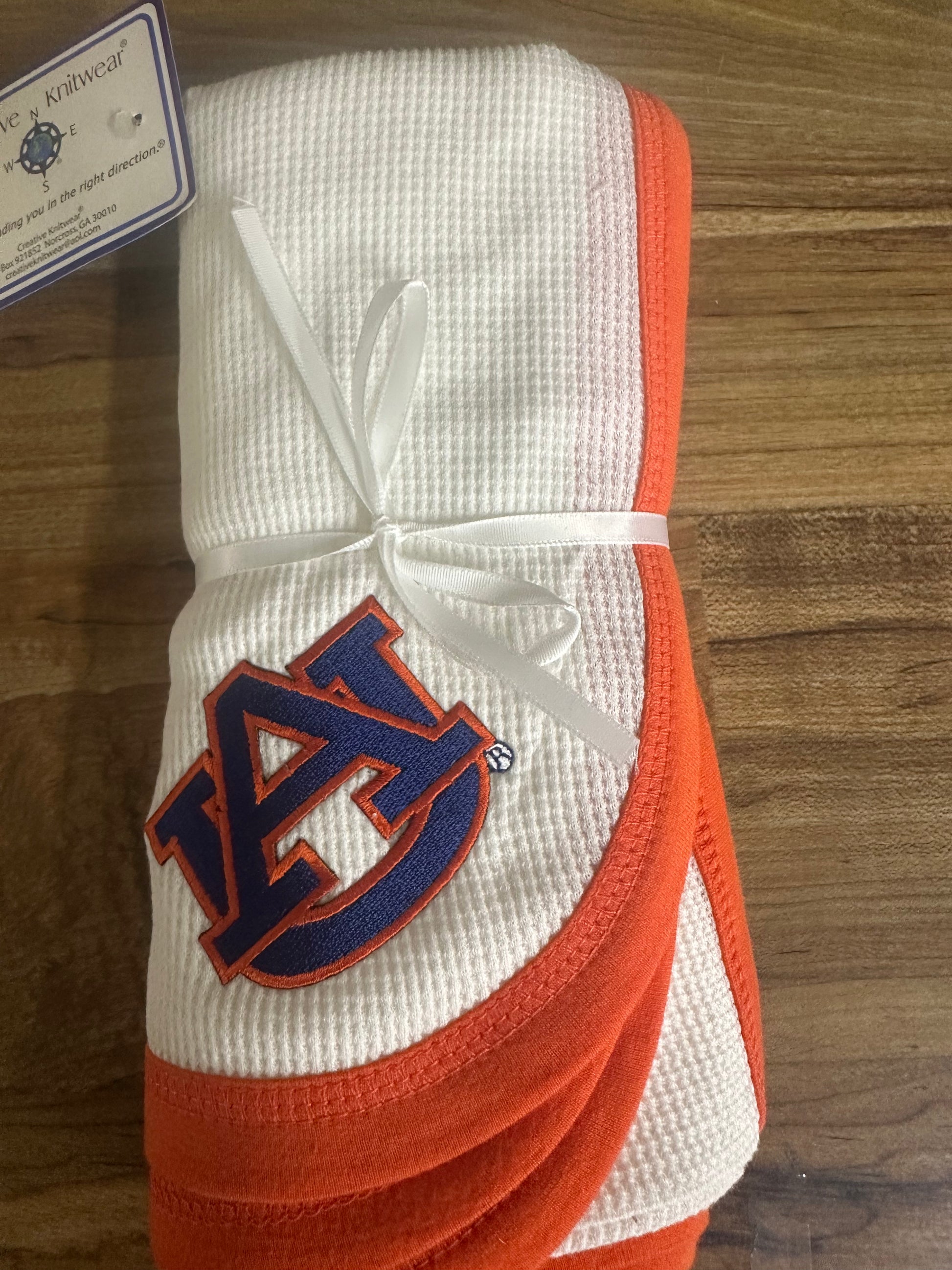 A folded white waffle-knit baby blanket with orange trim and a navy blue Auburn Tigers logo, tied with a white ribbon on a wooden surface.