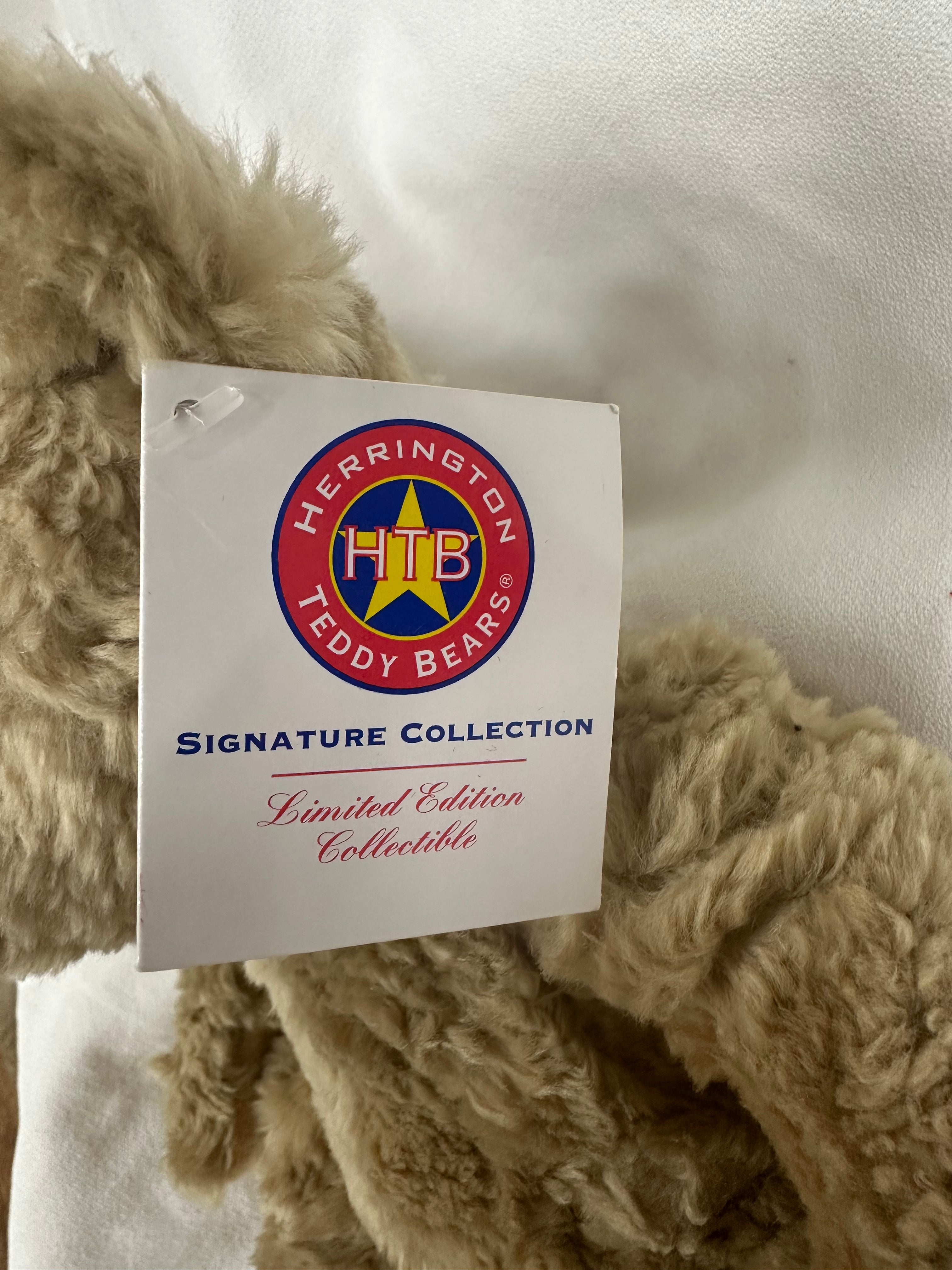 A close-up of a beige curly-fleece teddy bear with a white hangtag from Herrington Teddy Bears Signature Collection, set against a white fabric background.
