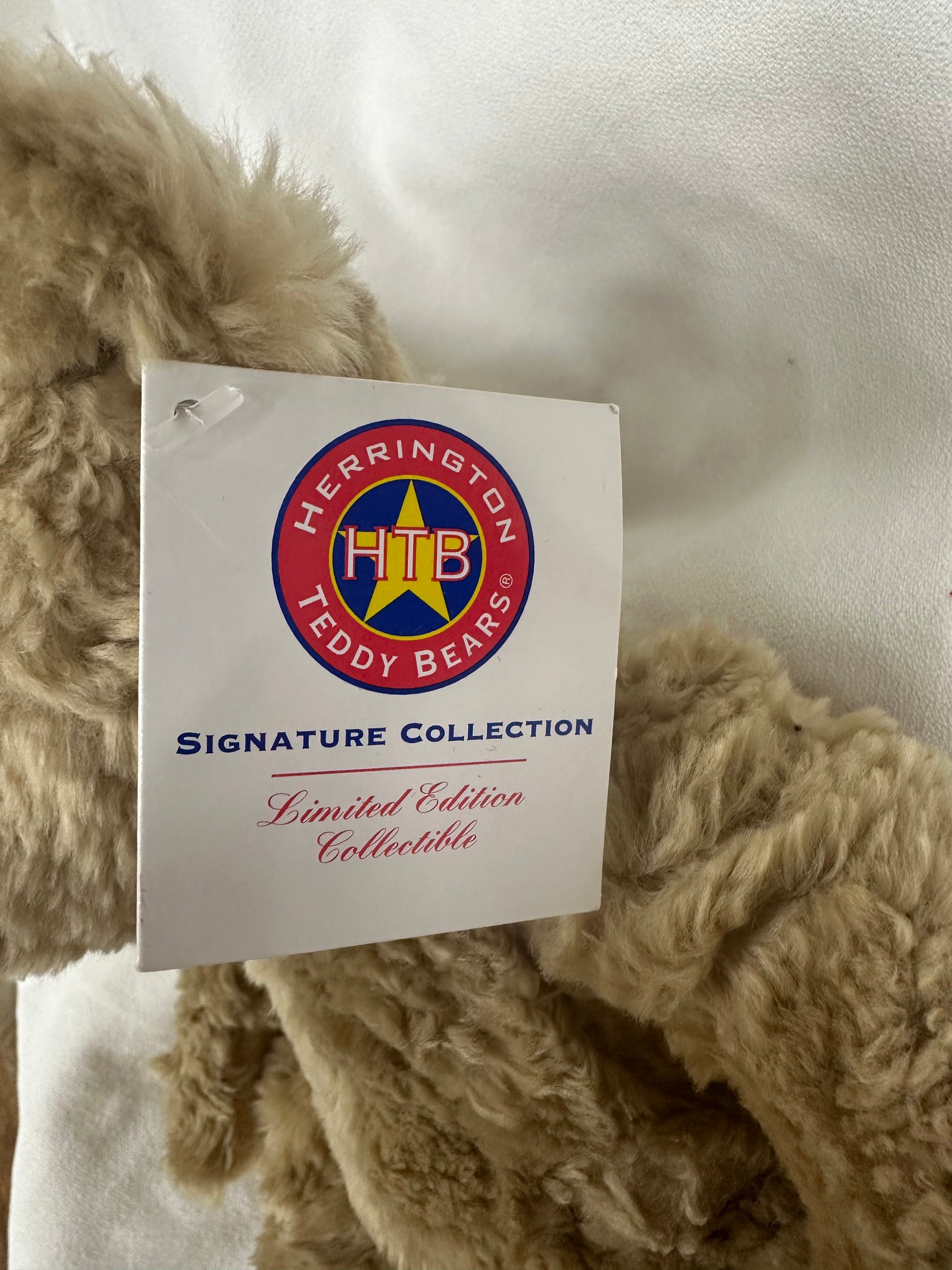 A close-up of a beige curly-fleece teddy bear with a white hangtag from Herrington Teddy Bears Signature Collection, set against a white fabric background.