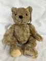 A classic Herringbone teddy bear plush toy in light brown, sitting on a white fabric background. The bear has black bead eyes, a black stitched nose, and a visible brand tag on its left foot reading 'HERRINGBONE TEDDY BEAR'.