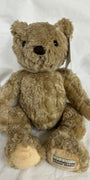 A classic Herringbone teddy bear plush toy in light brown, sitting on a white fabric background. The bear has black bead eyes, a black stitched nose, and a visible brand tag on its left foot reading 'HERRINGBONE TEDDY BEAR'.