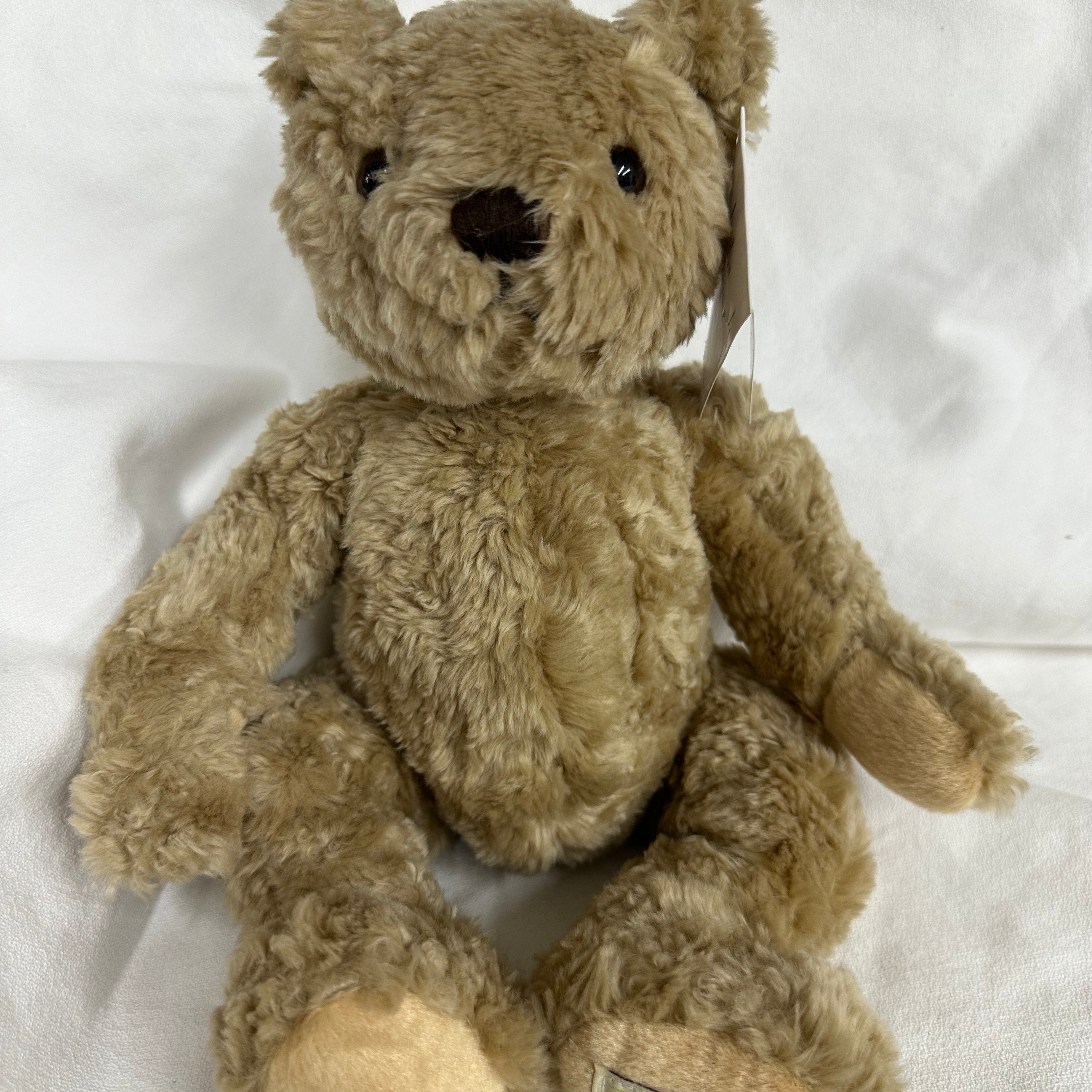 A classic Herringbone teddy bear plush toy in light brown, sitting on a white fabric background. The bear has black bead eyes, a black stitched nose, and a visible brand tag on its left foot reading 'HERRINGBONE TEDDY BEAR'.