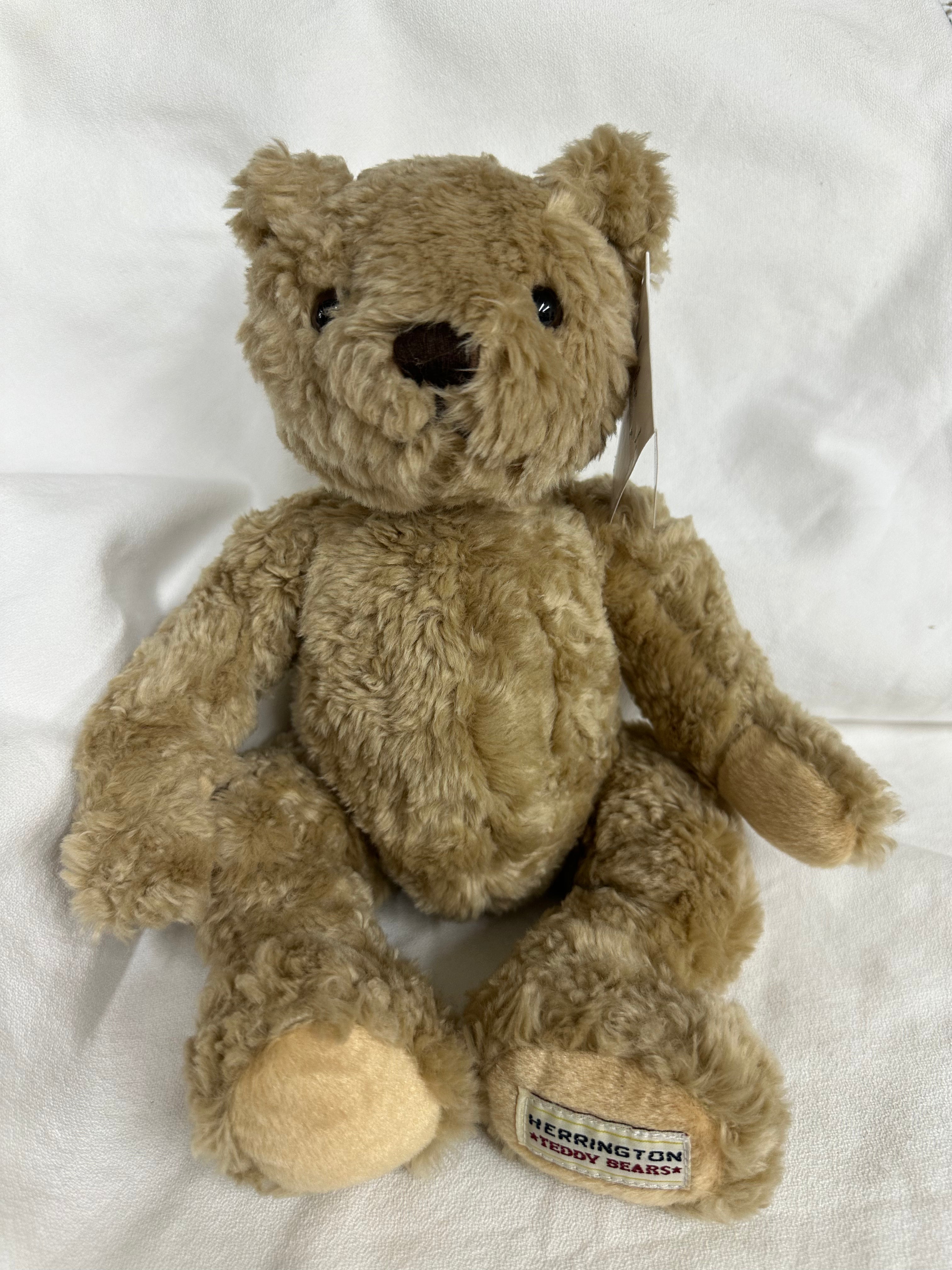 A classic Herringbone teddy bear plush toy in light brown, sitting on a white fabric background. The bear has black bead eyes, a black stitched nose, and a visible brand tag on its left foot reading 'HERRINGBONE TEDDY BEAR'.