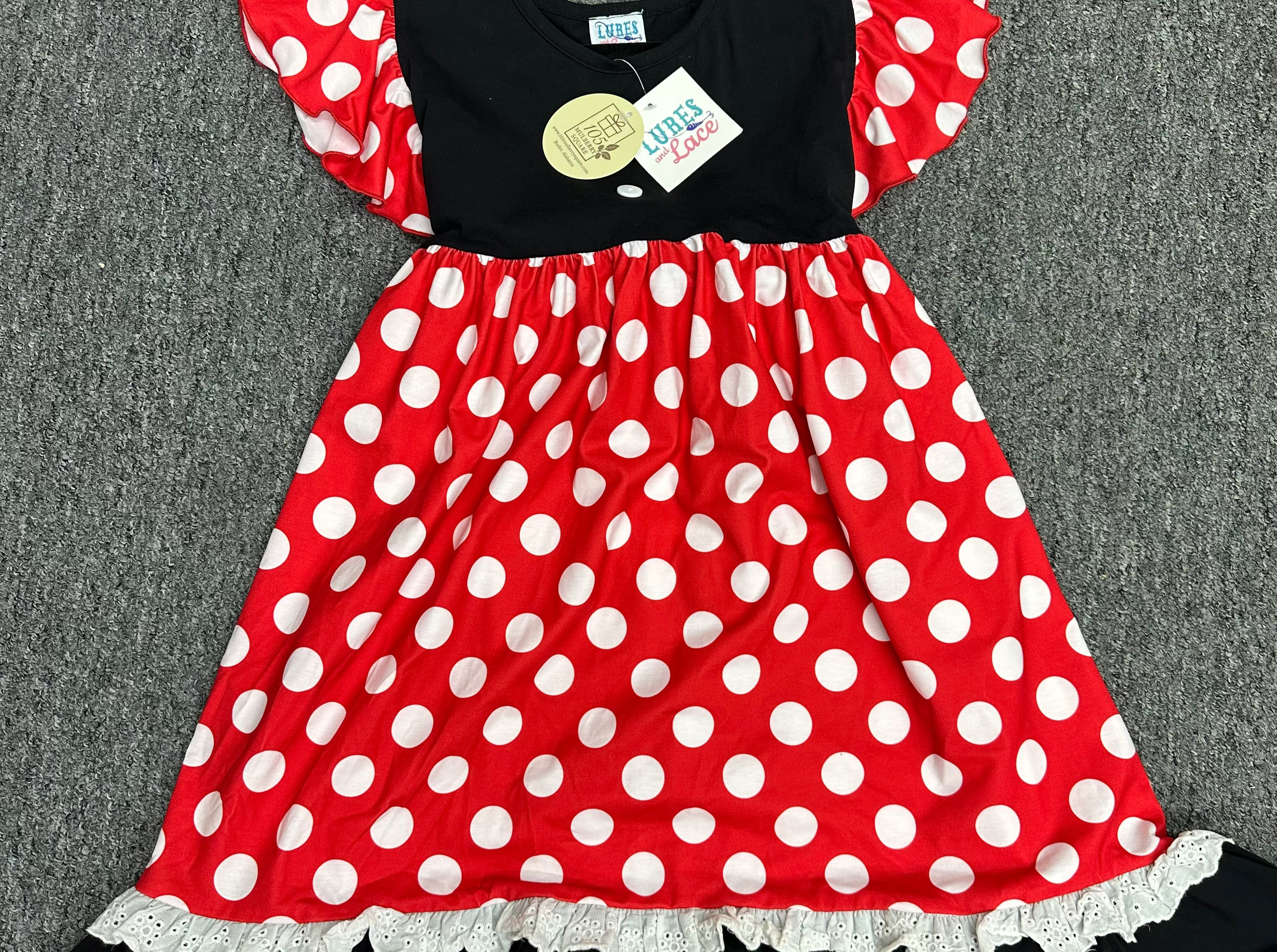 A children's dress laid flat on a gray textured surface, featuring a red and white polka-dot skirt, a black bodice, and matching red and white polka-dot ruffled shoulder straps.