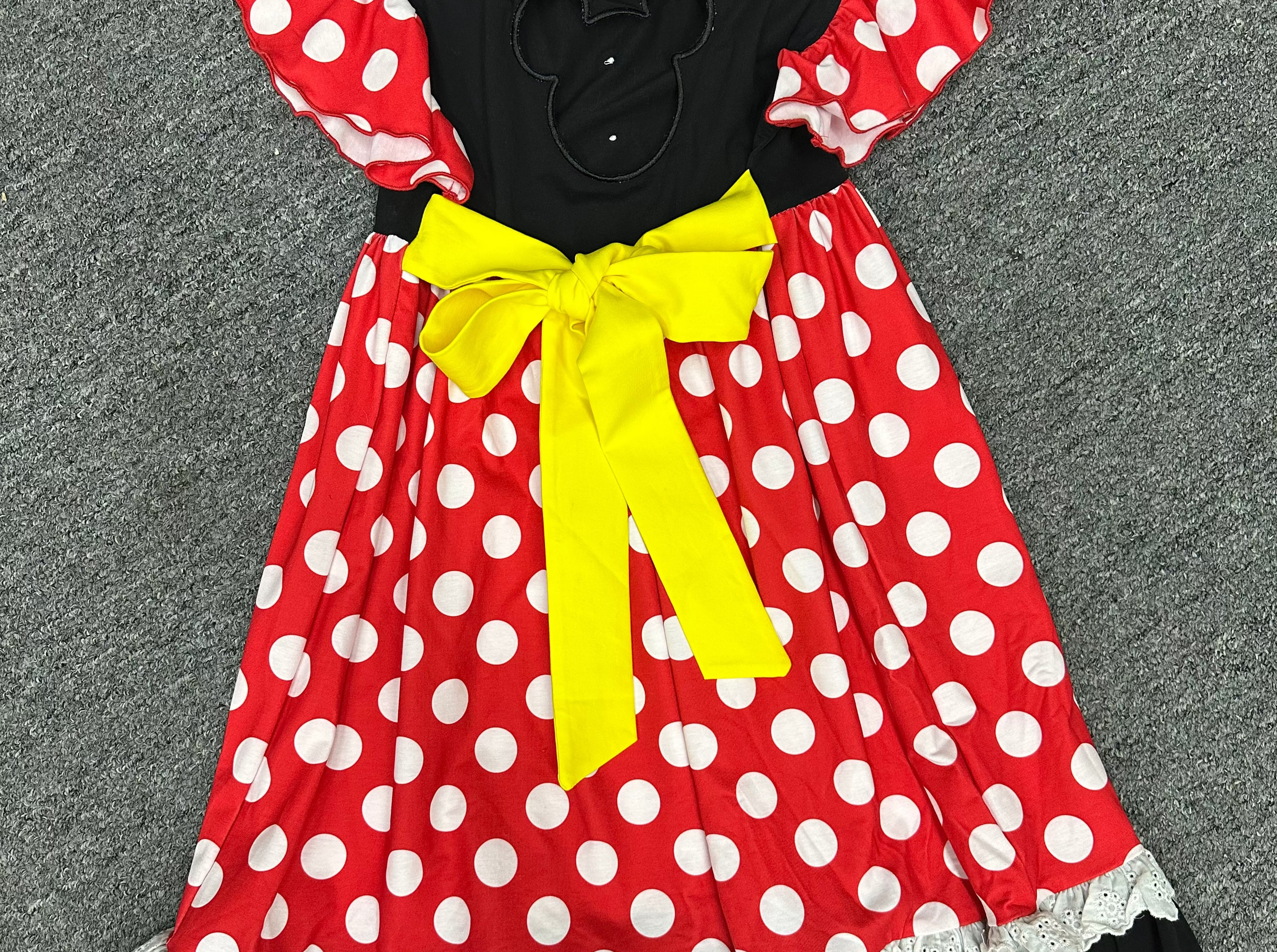 A Minnie Mouse-inspired dress laid flat on a gray carpet. The dress has a red base with white polka dots, black ruffled trim, a black bodice with a mouse-ear cutout, and a large yellow bow at the center.