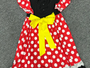 A Minnie Mouse-inspired dress laid flat on a gray carpet. The dress has a red base with white polka dots, black ruffled trim, a black bodice with a mouse-ear cutout, and a large yellow bow at the center.