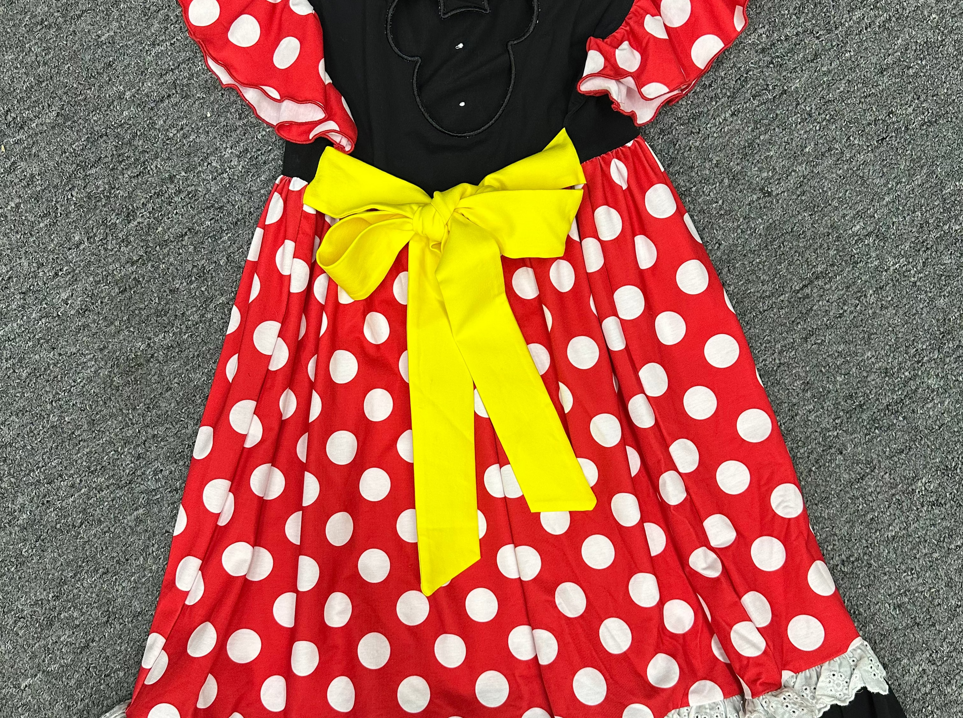 A Minnie Mouse-inspired dress laid flat on a gray carpet. The dress has a red base with white polka dots, black ruffled trim, a black bodice with a mouse-ear cutout, and a large yellow bow at the center.