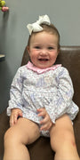 A baby girl sitting on a brown leather chair wearing a long-sleeve white and purple floral romper with a pink ruffled collar and a white bow in her hair.