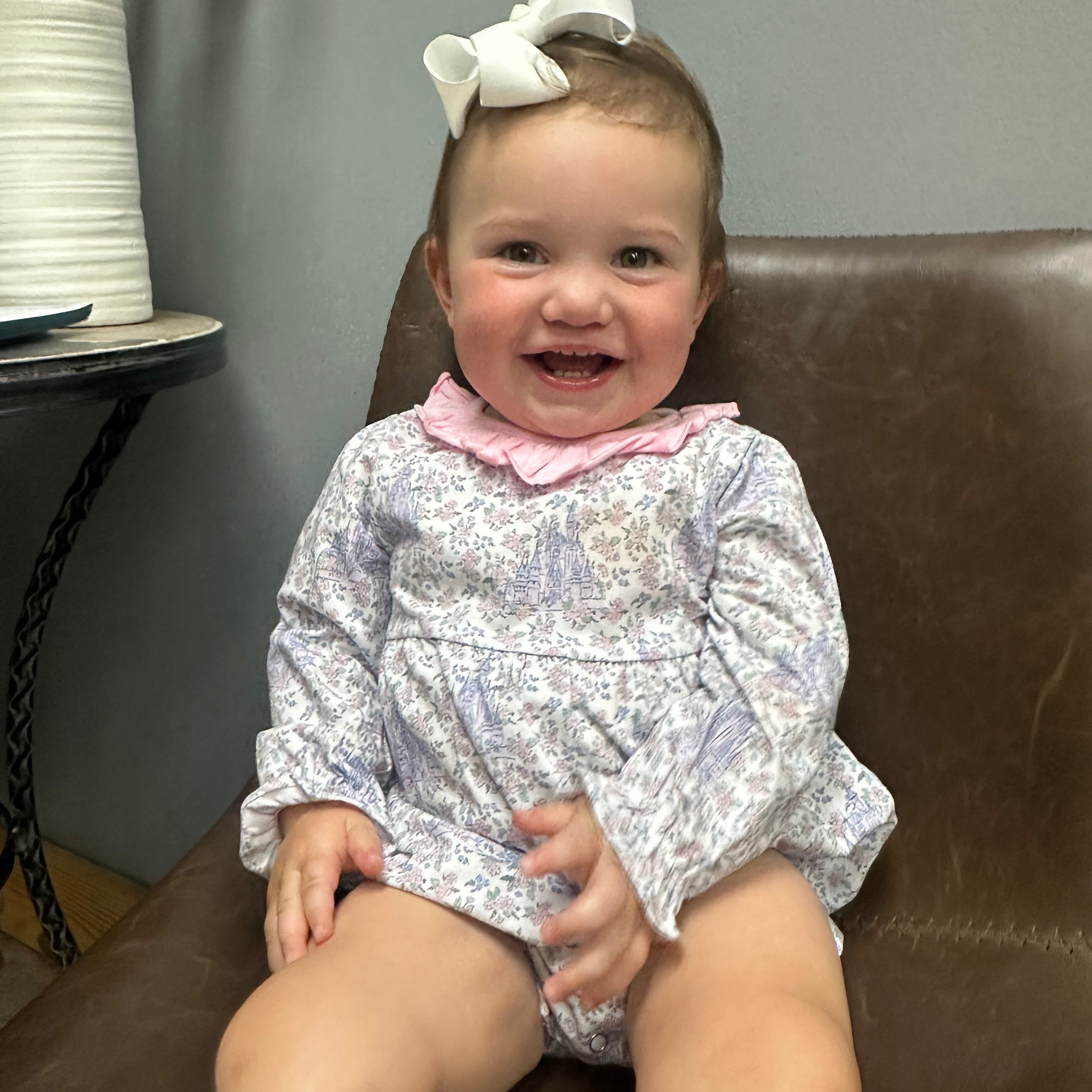 A baby girl sitting on a brown leather chair wearing a long-sleeve white and purple floral romper with a pink ruffled collar and a white bow in her hair.