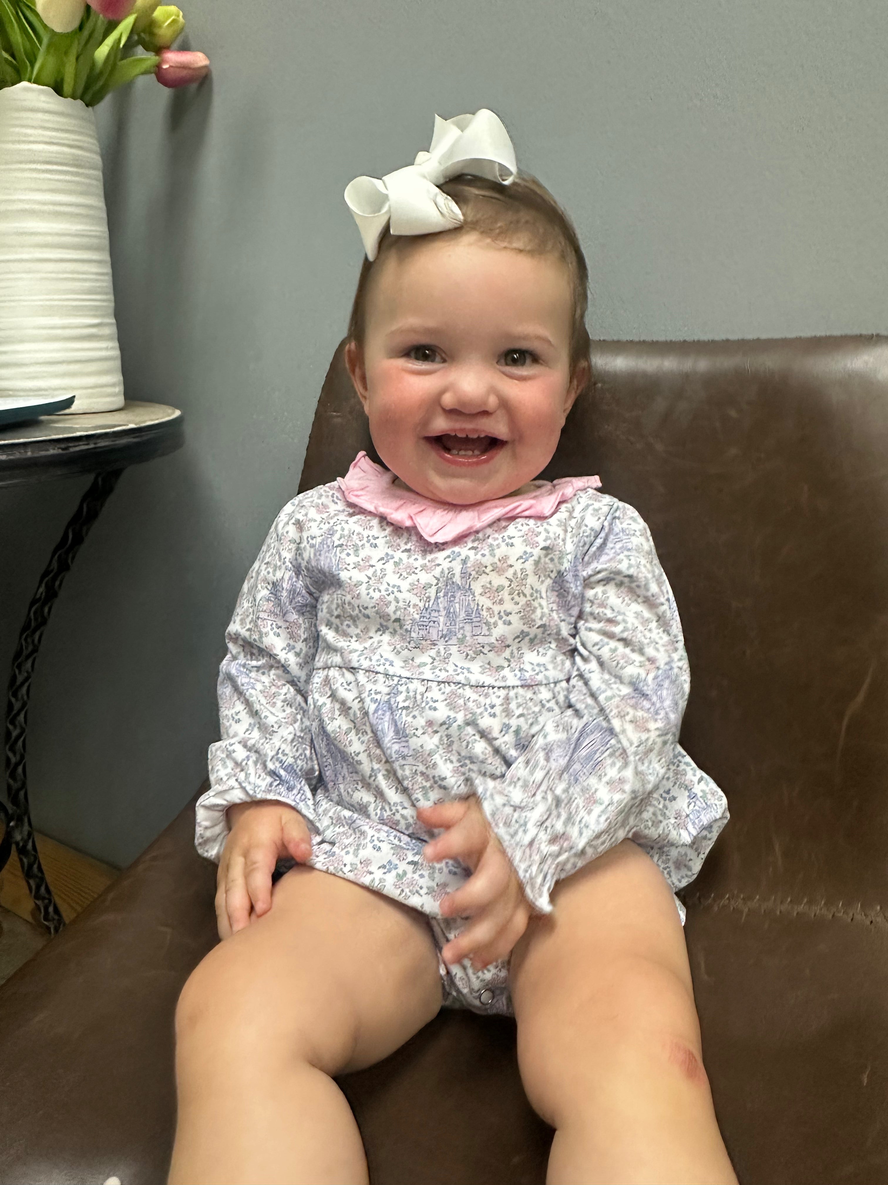 A baby girl sitting on a brown leather chair wearing a long-sleeve white and purple floral romper with a pink ruffled collar and a white bow in her hair.