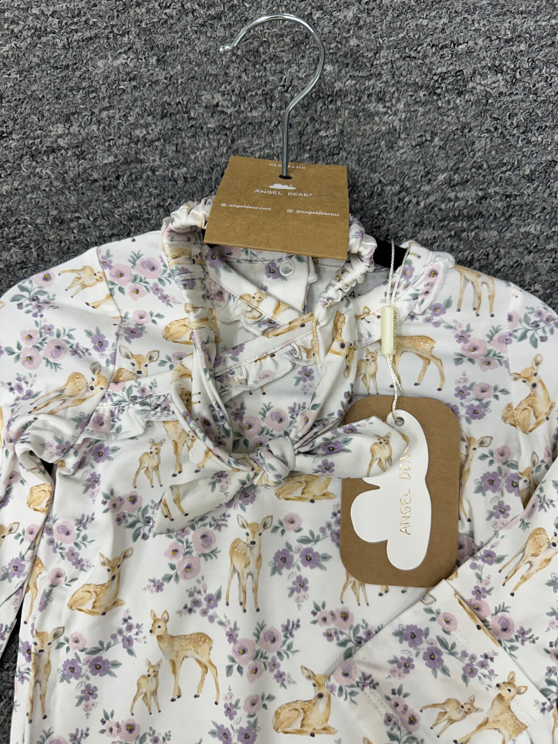 A long-sleeve baby top by Alice in Wonderland, featuring an all-over print of deer and purple flowers on a cream background. The shirt is displayed on a metal hanger with its original brown paper tags attached, set against a dark gray textured background.