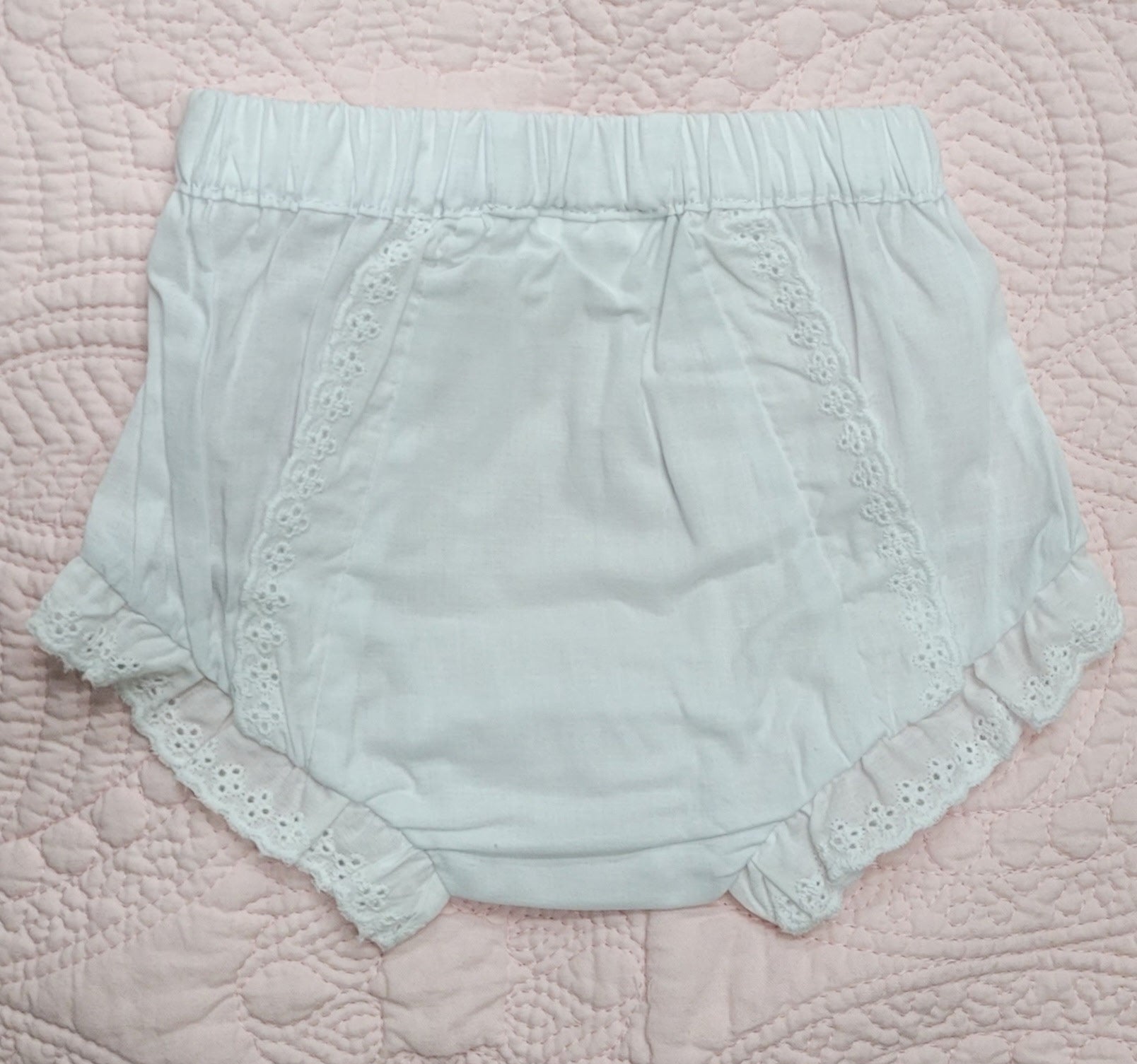 A pair of light mint green baby bloomers with eyelet lace trim, laid flat on a pink quilted blanket.