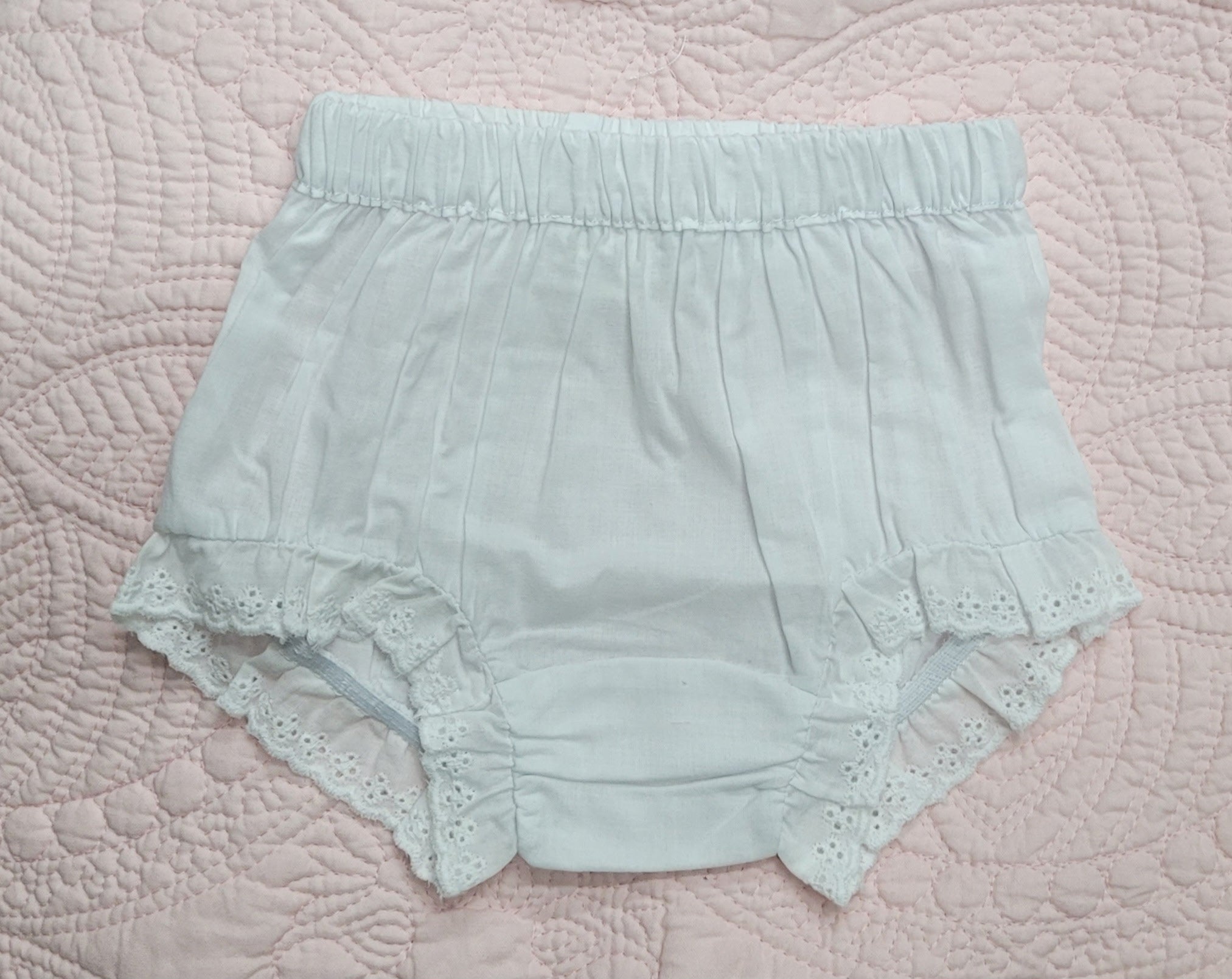 A pair of pale mint green baby bloomers with a gathered elastic waistband and eyelet lace trim, laid flat on a pink quilted blanket.