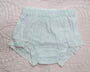 A pair of pale mint green baby bloomers with a gathered elastic waistband and eyelet lace trim, laid flat on a pink quilted blanket.