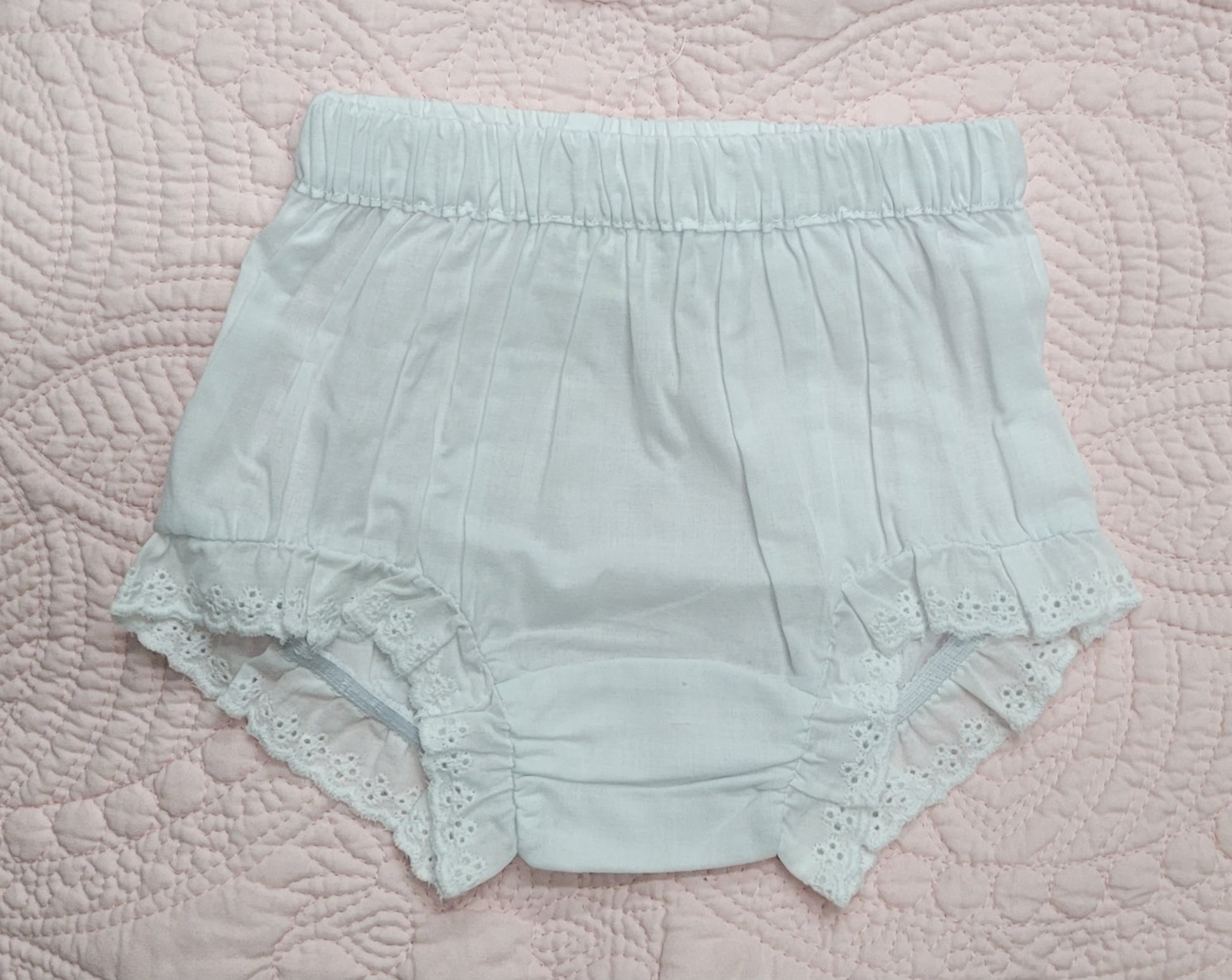 A pair of pale mint green baby bloomers with a gathered elastic waistband and eyelet lace trim, laid flat on a pink quilted blanket.
