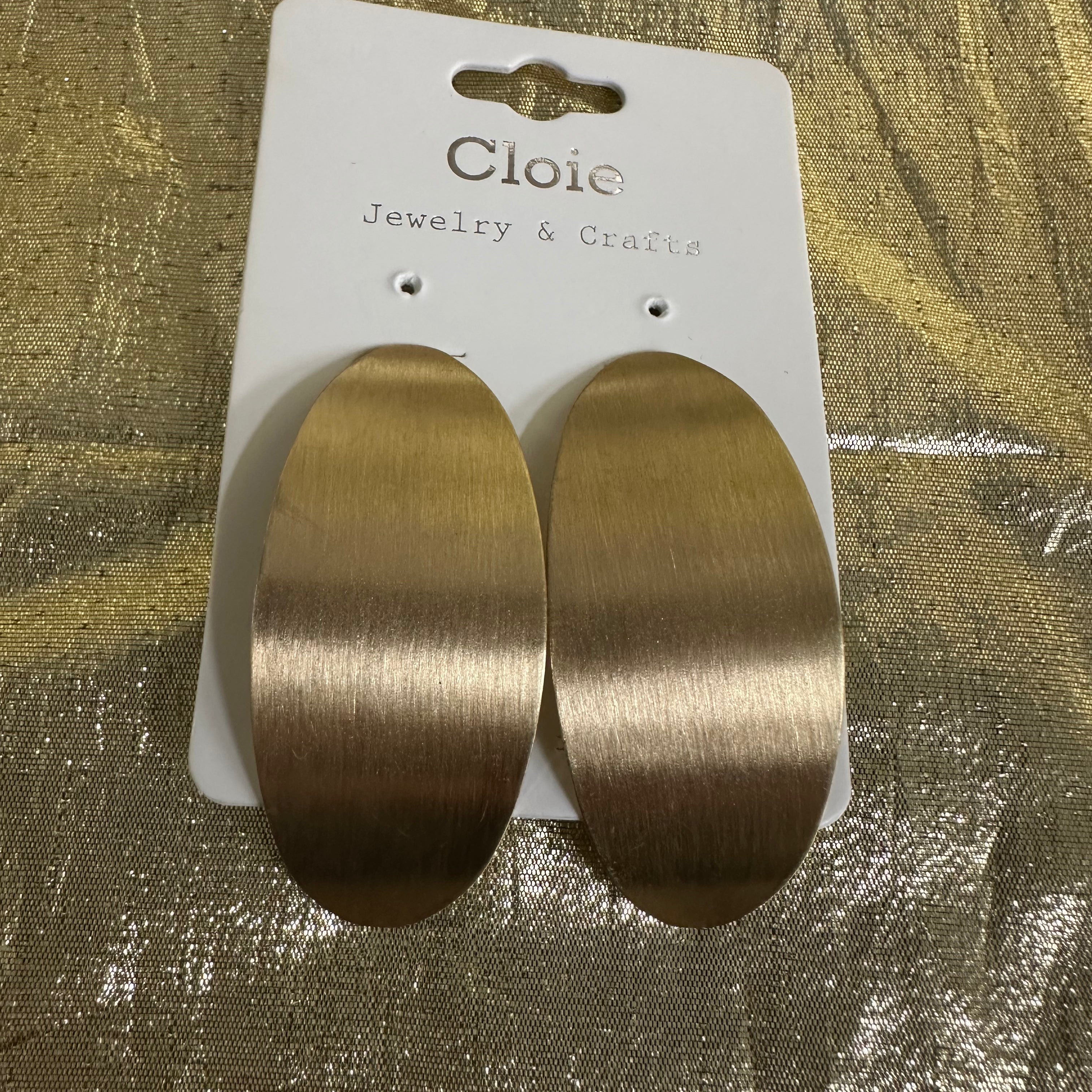 A pair of large oval drop earrings with a brushed gold finish, attached to a white display card labeled 'Cloie Jewelry & Crafts' and placed on a textured gold surface.