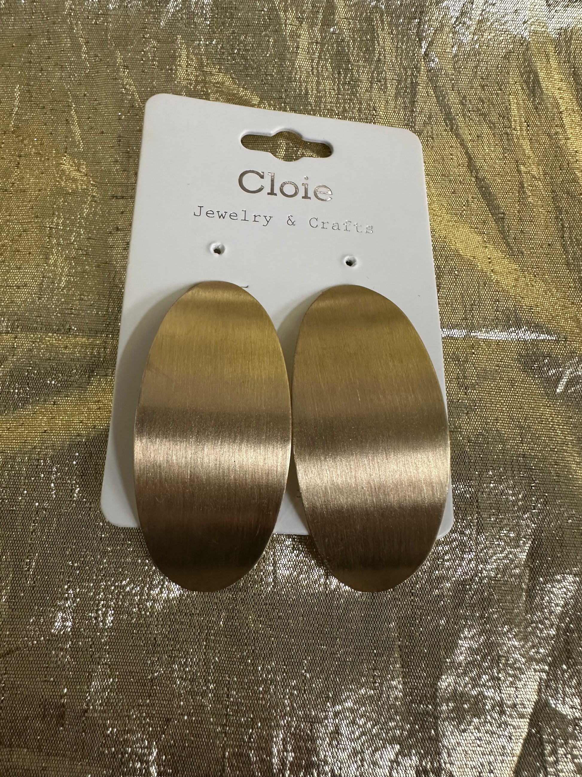 A pair of large oval drop earrings with a brushed gold finish, attached to a white display card labeled 'Cloie Jewelry & Crafts' and placed on a textured gold surface.