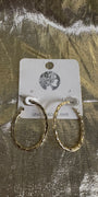 A pair of textured gold hoop earrings displayed on a white cardboard backing card, set against a shiny, crinkled metallic gold background.