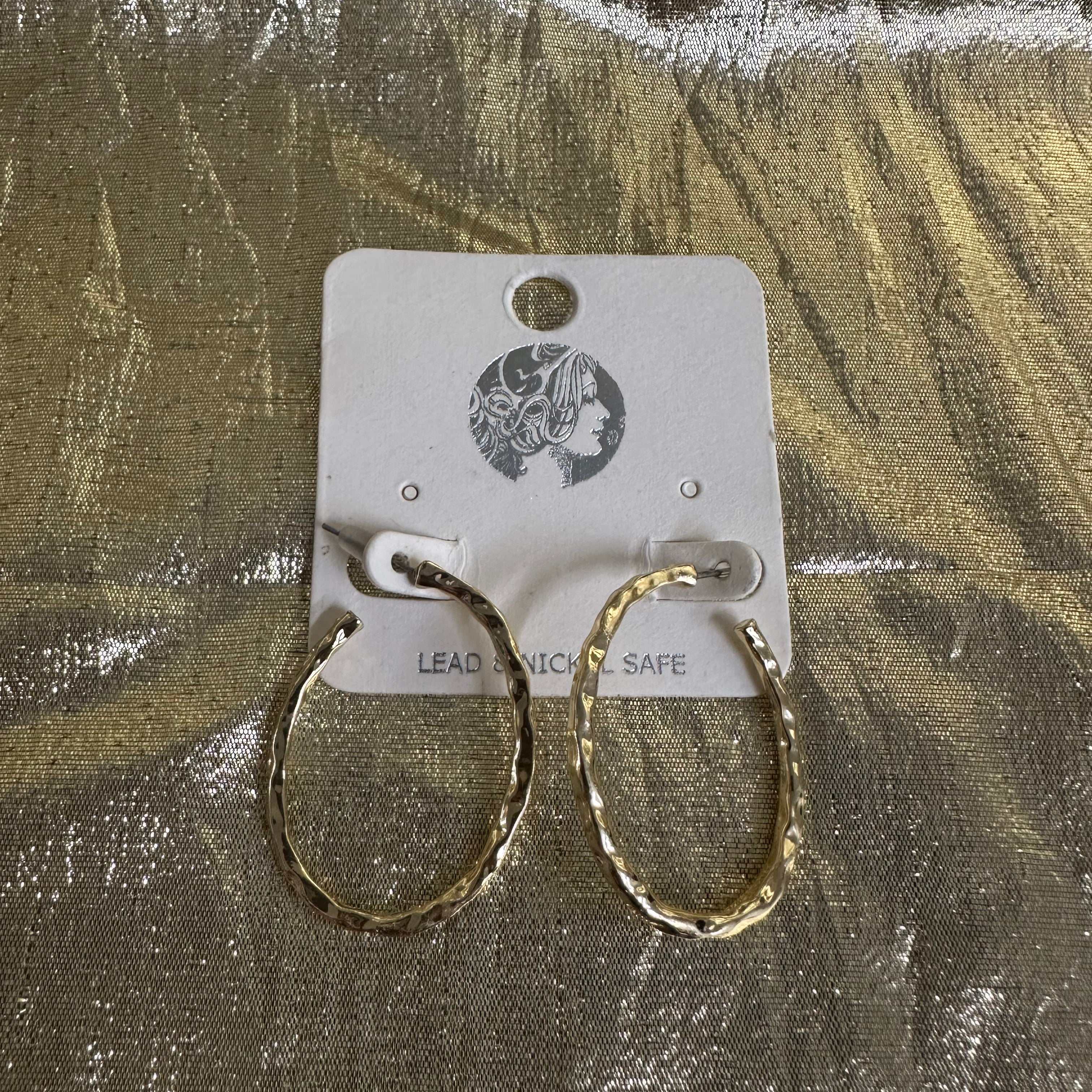 A pair of textured gold hoop earrings displayed on a white cardboard backing card, set against a shiny, crinkled metallic gold background.