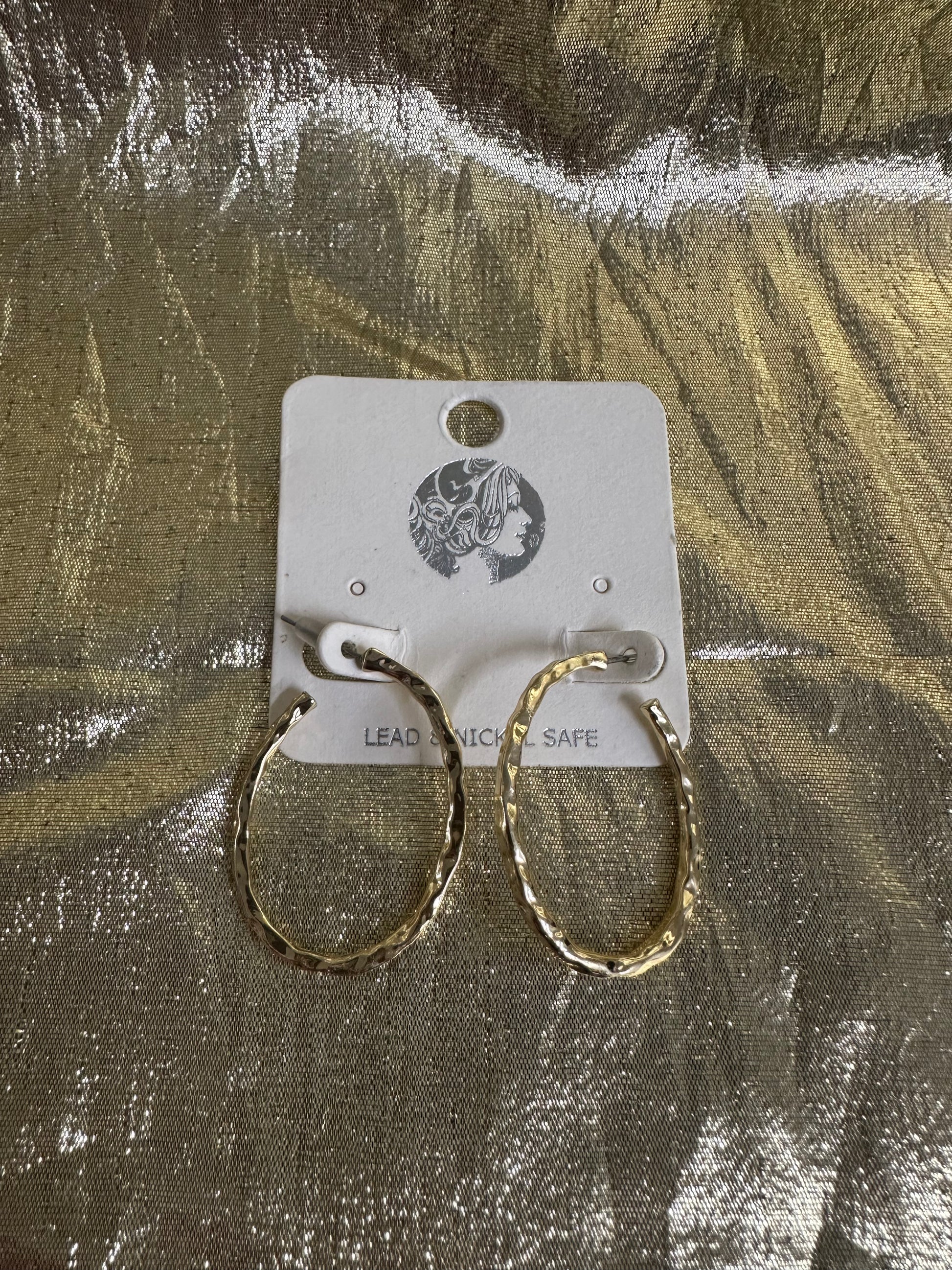A pair of textured gold hoop earrings displayed on a white cardboard backing card, set against a shiny, crinkled metallic gold background.