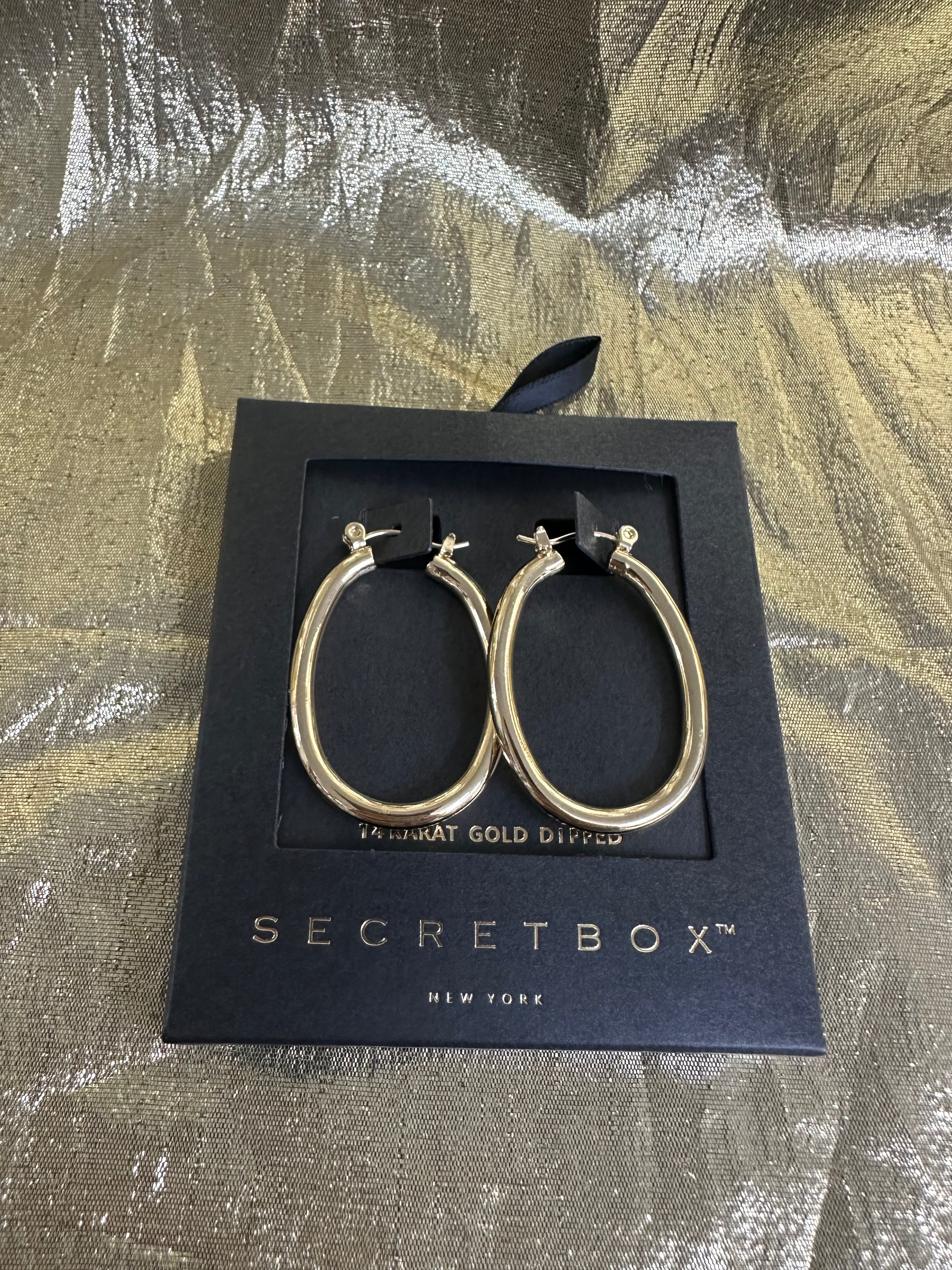 A pair of gold hoop earrings displayed on a black Secretbox New York gift box, set against a textured metallic gold background.