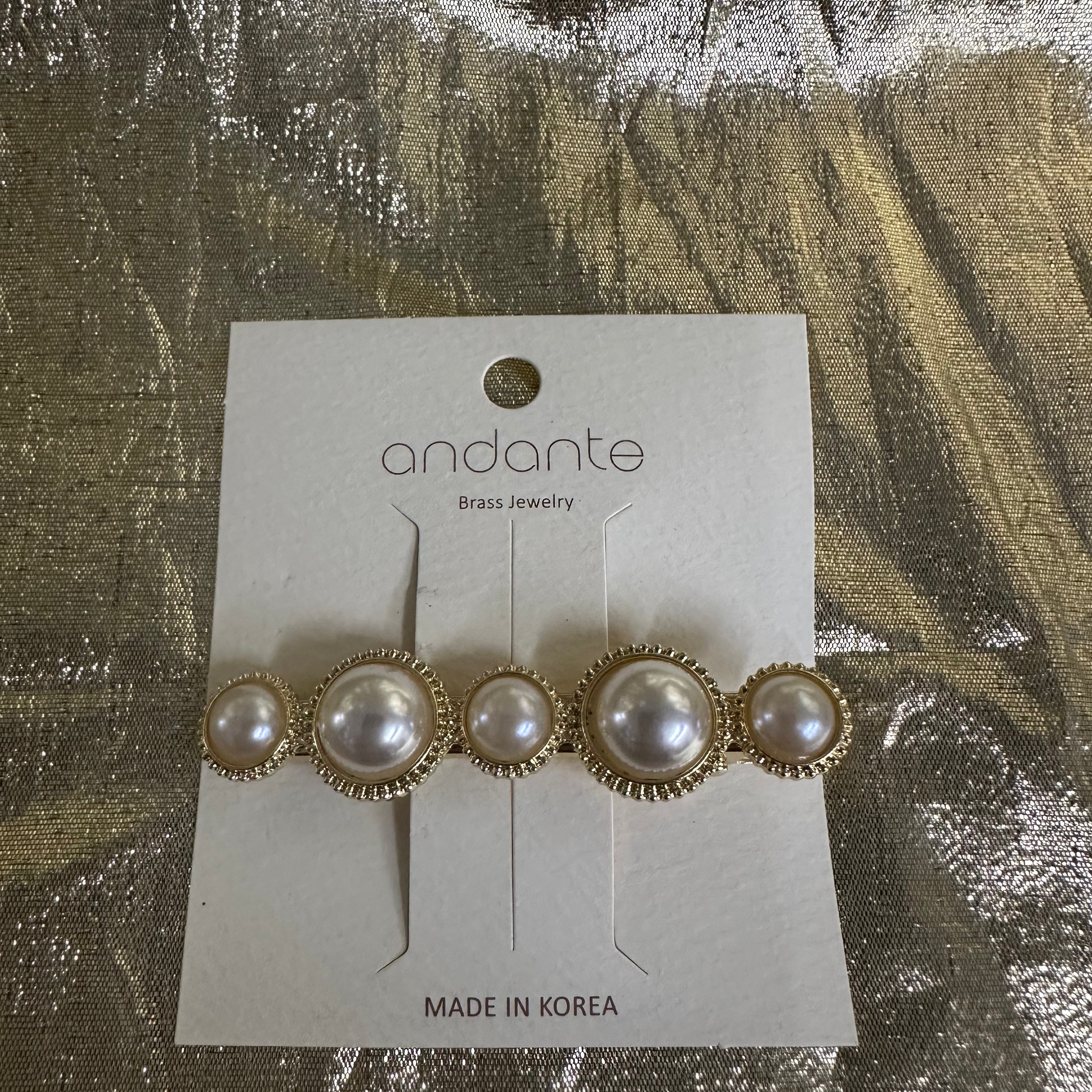 Andante gold and pearl hair clip on a white display card, photographed on a textured gold foil background.