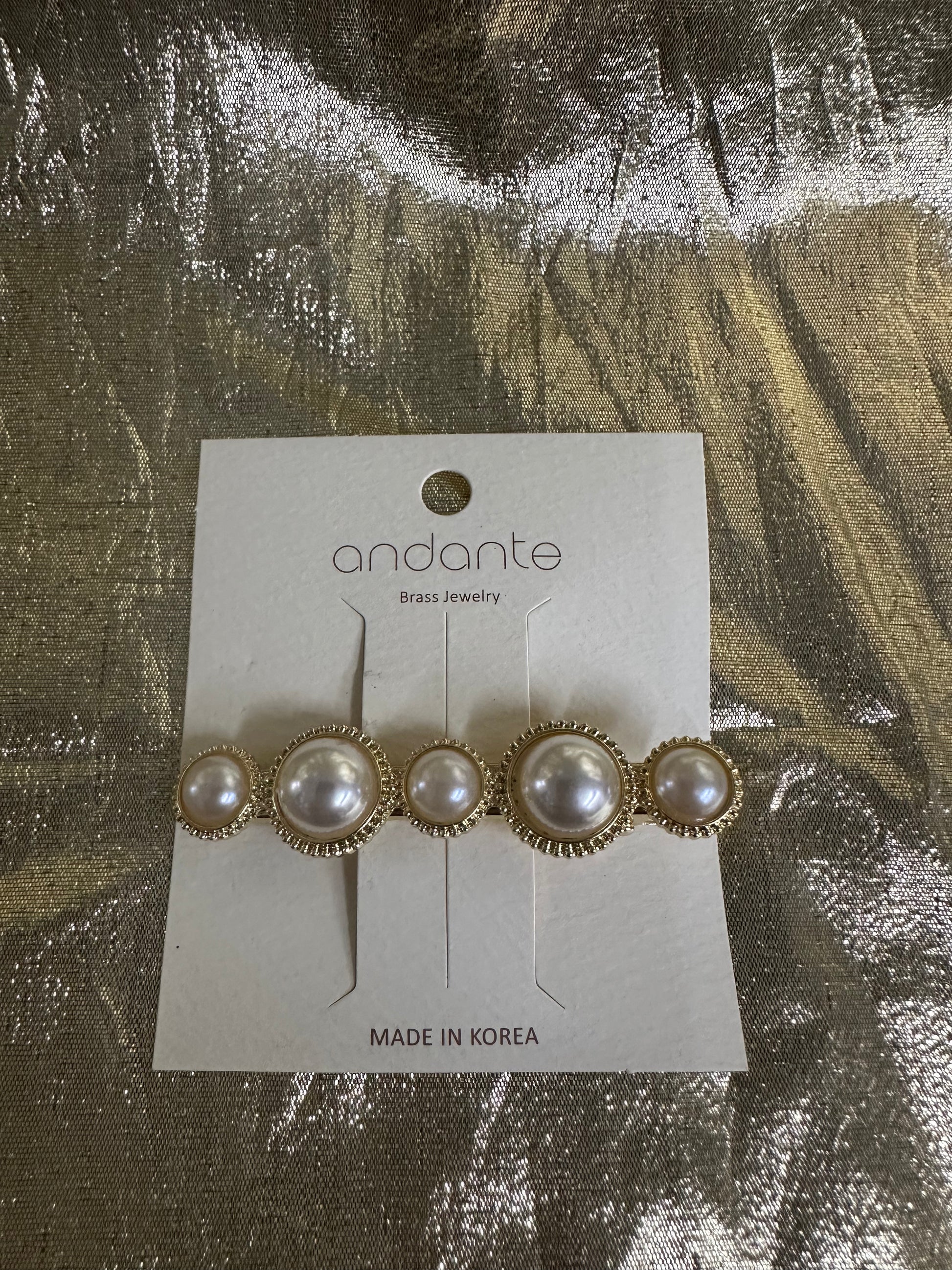 Andante gold and pearl hair clip on a white display card, photographed on a textured gold foil background.