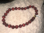 A beaded necklace made of glossy dark red round beads with a small gold bell accent, resting on a white textured blanket.