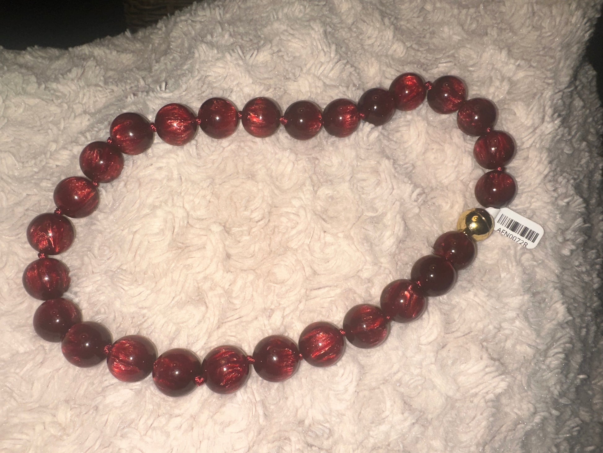 A beaded necklace made of glossy dark red round beads with a small gold bell accent, resting on a white textured blanket.