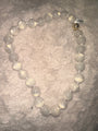 A beaded necklace made of large, smooth, milky white oval beads with a small metallic gold accent bead, displayed on a textured, light beige faux fur surface.
